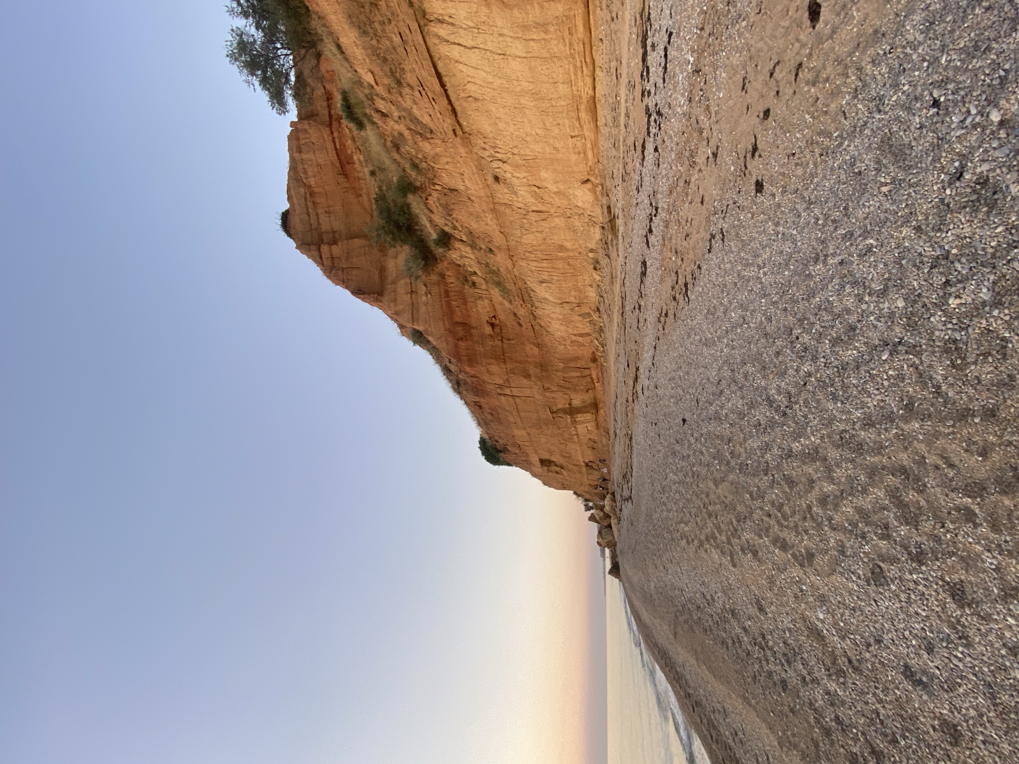 This image shows a view of the coast in Sevastopol, Ukraine, at what appears to be sunrise or sunset. A tall, reddish-orange cliff face dominates the right side of the frame, its layered texture suggesting sedimentary rock. Sparse green vegetation grows on top of and along the side of the cliff. The cliff meets a pebble and sand beach that stretches to the left. Small waves gently lap at the shore on the far left. The sky is a clear, pale blue, transitioning to a lighter, almost white hue near the horizon. A few small figures can be seen walking along the beach near the base of the cliff, indicating human presence in the scene. The overall atmosphere is serene and natural.