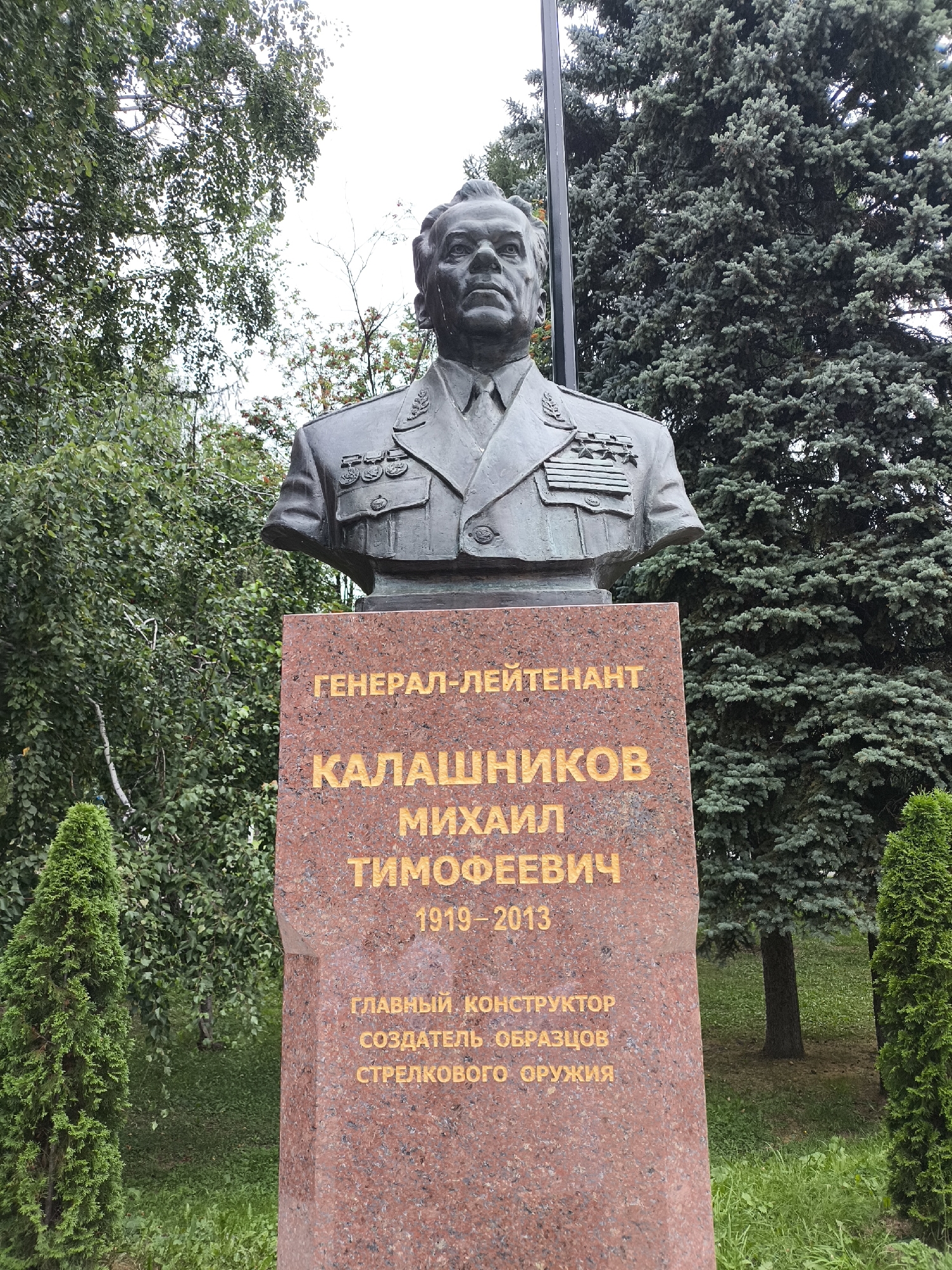 A bust of Mikhail Kalashnikov, a Soviet and Russian general, designer, and inventor, is mounted on a red granite pedestal. The pedestal has gold lettering that reads "GENERAL-LIEUTENANT KALASHNIKOV MIKHAIL TIMOFEEVICH 1919-2013", followed by "CHIEF DESIGNER CREATOR OF SMALL ARMS MODELS". The bust is depicted wearing a military uniform with multiple medals. The scene is outdoors, with lush green trees and grass surrounding the monument. A tall, dark pole is visible behind the bust, likely a flagpole. This monument is located in Izhevsk, Russia, at the Kalashnikov Concern museum.