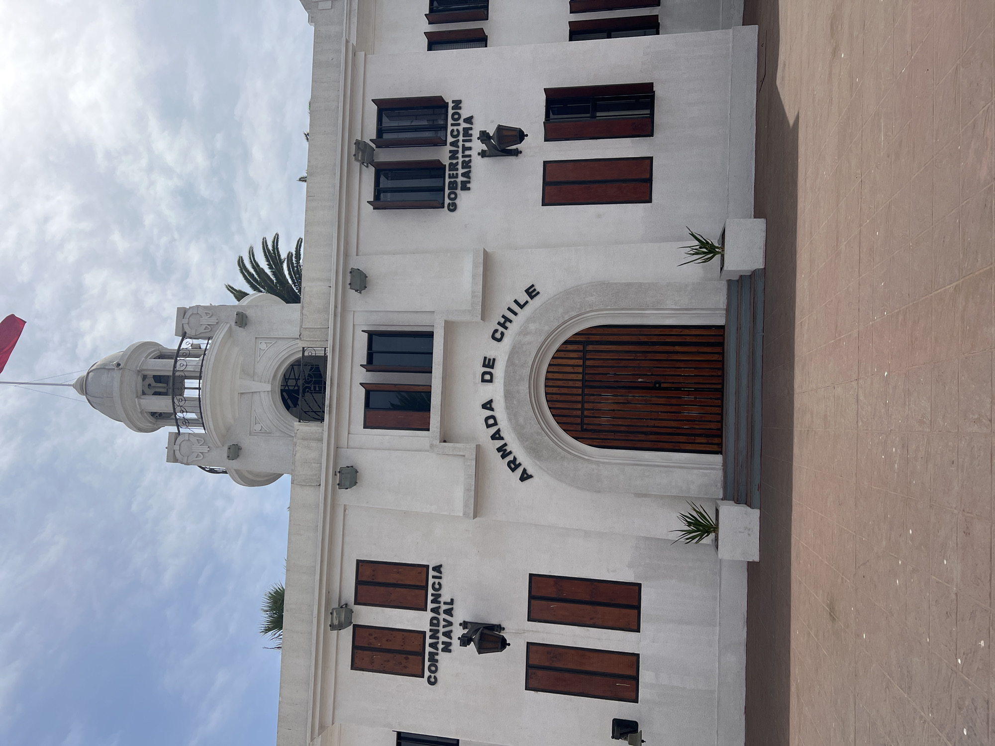 The image shows a white building, likely a government building, in Arica, Chile. "GOBERNACION MARITIMA" and "ARMADA DE CHILE" are inscribed on the facade. Below that, the words "COMANDANCIA NAVAL" are displayed. The building has several rectangular windows with dark-colored shutters. The main entrance is an arched wooden door with bars. Two small plants flank the entrance. The building's design includes a tower-like structure on the left side. The plaza flooring is visible in the bottom right corner. The sky above the building is cloudy.