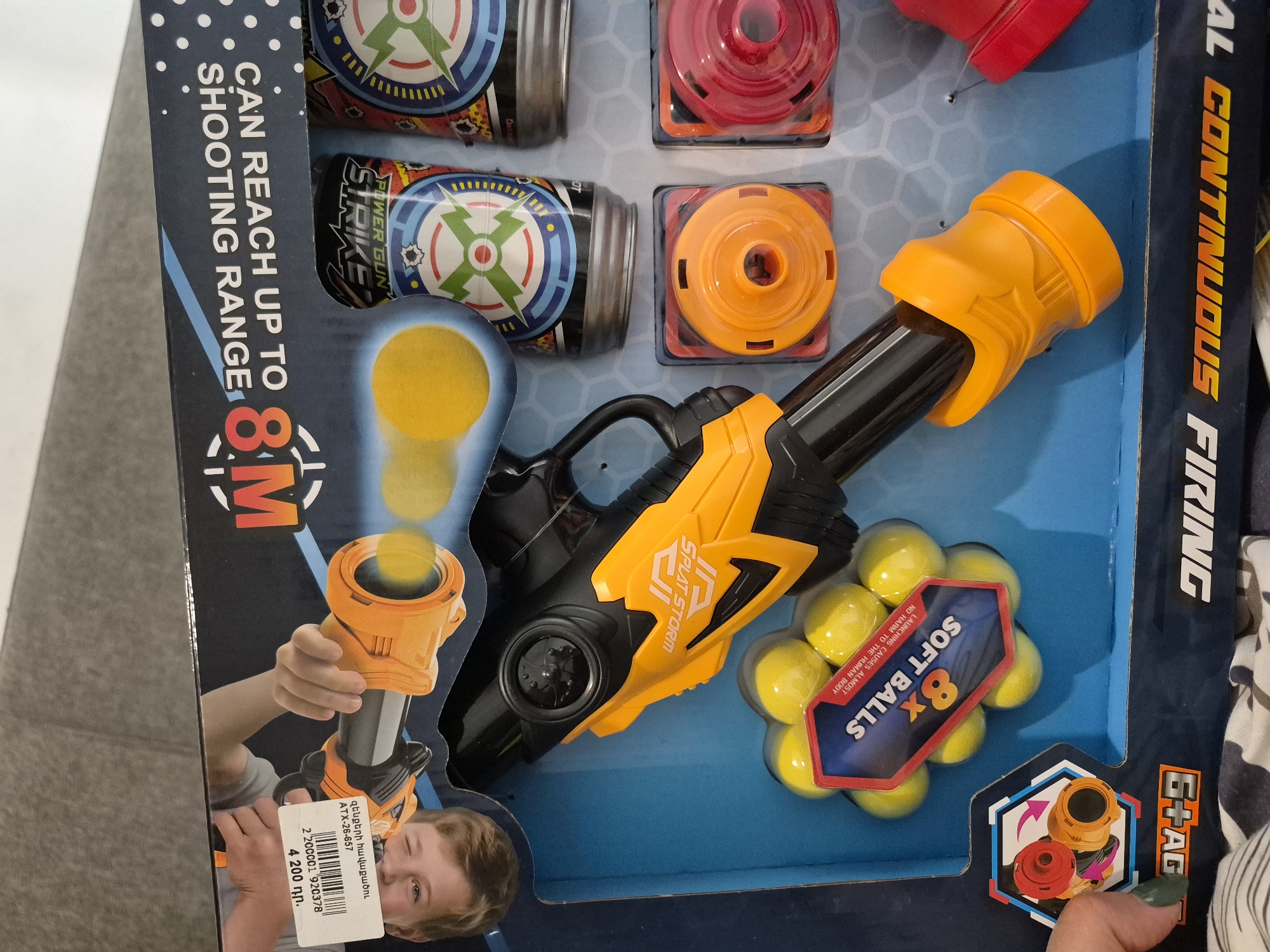 The image displays a toy gun set, likely a foam or soft ball blaster, presented in its retail packaging. The scene appears to be indoors, possibly in a store in Yerevan, Armenia, as indicated by the price tag.
The main subject is a black and yellow toy gun, branded "SPLAT STORM". Alongside it, the set includes eight yellow "SOFT BALLS," with packaging text stating "LAUNCHING CAUSES ALMOST NO HARM TO THE HUMAN BODY." Also included are four targets: two cylindrical targets featuring a "POWER GUN STRIKE" logo, and two square-based targets with round tops, one red and one orange.
The packaging highlights key features: "CAN REACH UP TO 8M SHOOTING RANGE" and partially visible "CONTINUOUS FIRING." An age recommendation of "6+ AGE" is also visible. The box art depicts a young boy winking and holding a similar toy gun, suggesting a playful experience. A hand with painted fingernails is visible in the bottom right corner, partially interacting with the packaging.
A price tag sticker is affixed to the package, displaying Armenian text: "զենքերի հավաքածու" (weapon set), product code "ATX-26-657," a barcode number "2 200001 920378," and the price "4 200 դր." (4,200 Armenian dram).