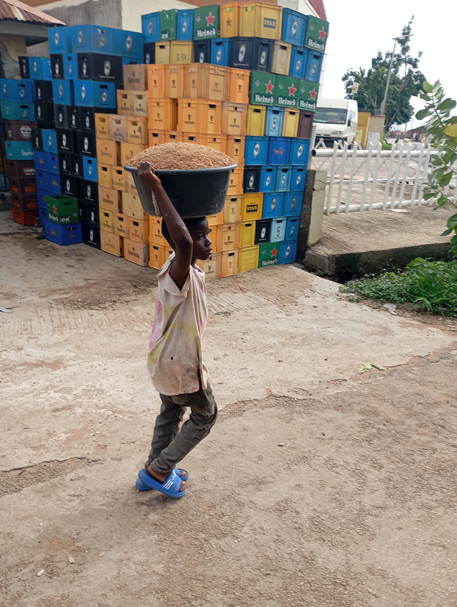 In the city of Shendam, Nigeria, a young boy walks across a dusty, uneven dirt road, carrying a large basin filled with grain balanced on his head. He is wearing a faded, light-colored t-shirt with pink and yellow tie-dye accents and dark pants. On his feet are bright blue slip-on sandals. He is walking from left to right across the frame, looking slightly to his right. Behind him, to his left, is a very large stack of colorful plastic crates, predominantly blue and orange, with some black and green crates mixed in. Many of these crates bear logos such as "Heineken" and "STAR". To the right of the boy, further back, is a white fence and a glimpse of a white van. The environment appears to be an outdoor, possibly commercial, area with evidence of storage and transport.
