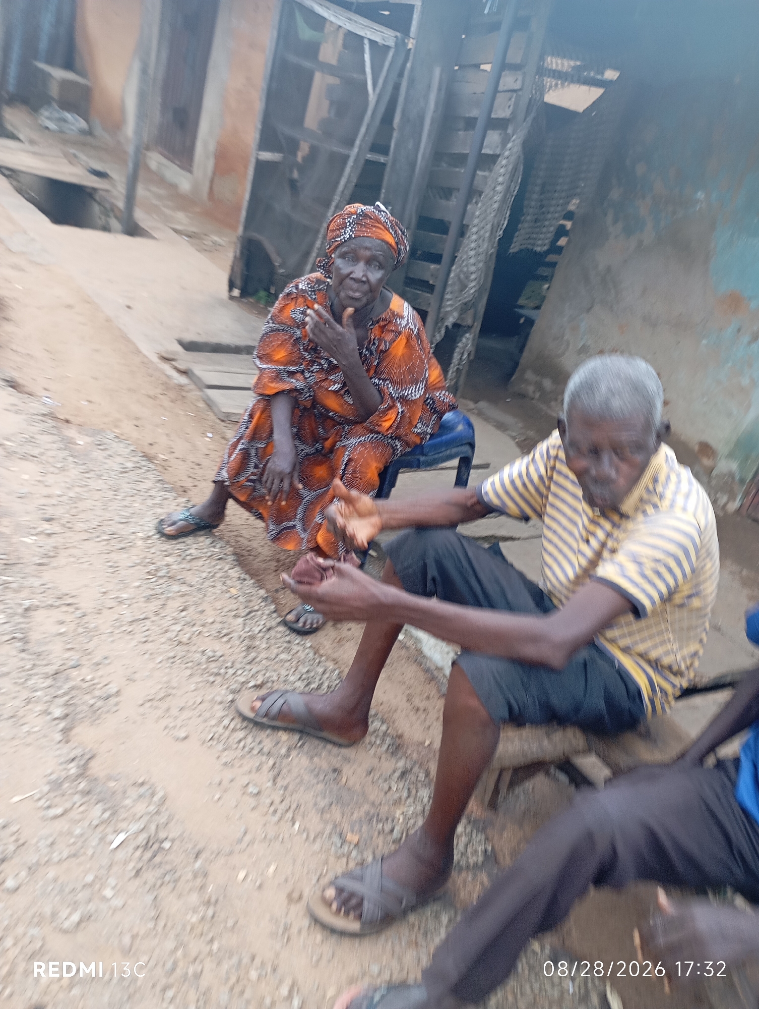 In Shendam, Nigeria, two elderly individuals are seated outdoors in what appears to be a village setting. To the left, an older woman with gray hair wears an orange and black patterned dress and a matching headscarf. She is seated on a blue chair, resting her hand near her chin. To her right, an older man with gray hair and dark skin is seated on a wooden bench. He is wearing a yellow and blue striped short-sleeved shirt and black shorts. His hands are visible, one resting on his leg and the other in his lap. The ground is covered with sand and small stones, and wooden planks are visible in the background, suggesting a somewhat rustic environment. A dilapidated building with a light blue painted section is also visible behind them, as well as wooden stairs. The overall scene depicts a moment of rest or conversation in a rural Nigerian locale.