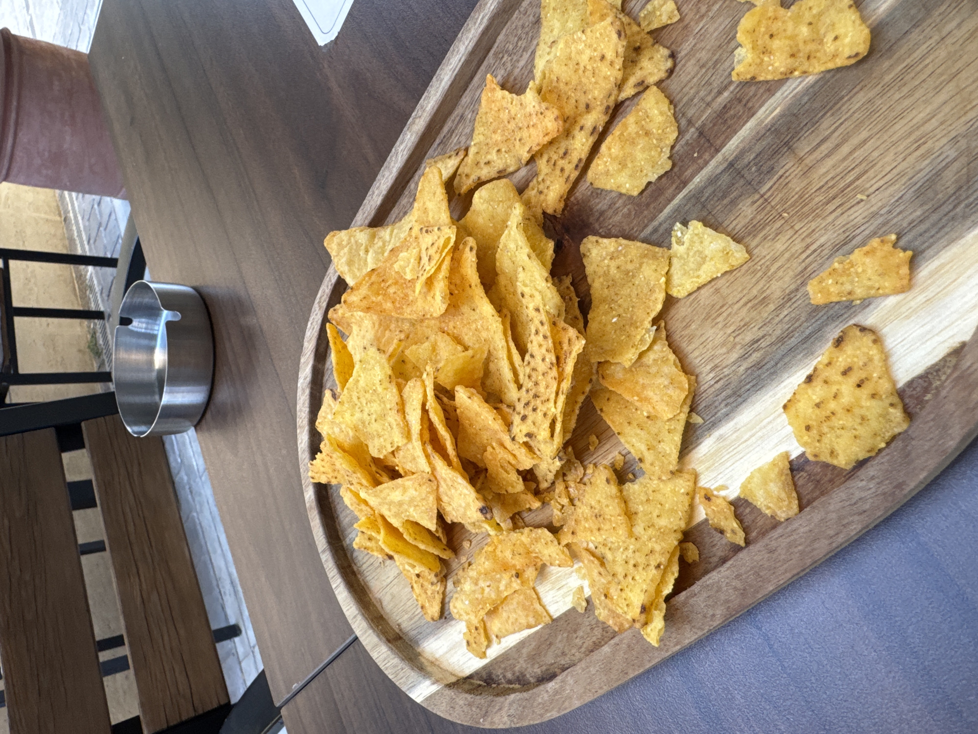 A wooden platter filled with a pile of golden tortilla chips sits on a dark wooden table. Some chips are broken into smaller pieces. In the background, a silver ashtray is visible on the table, and beyond that, a wooden bench and a paved outdoor area suggest a cafe or restaurant setting, possibly in Podgorica, Montenegro.