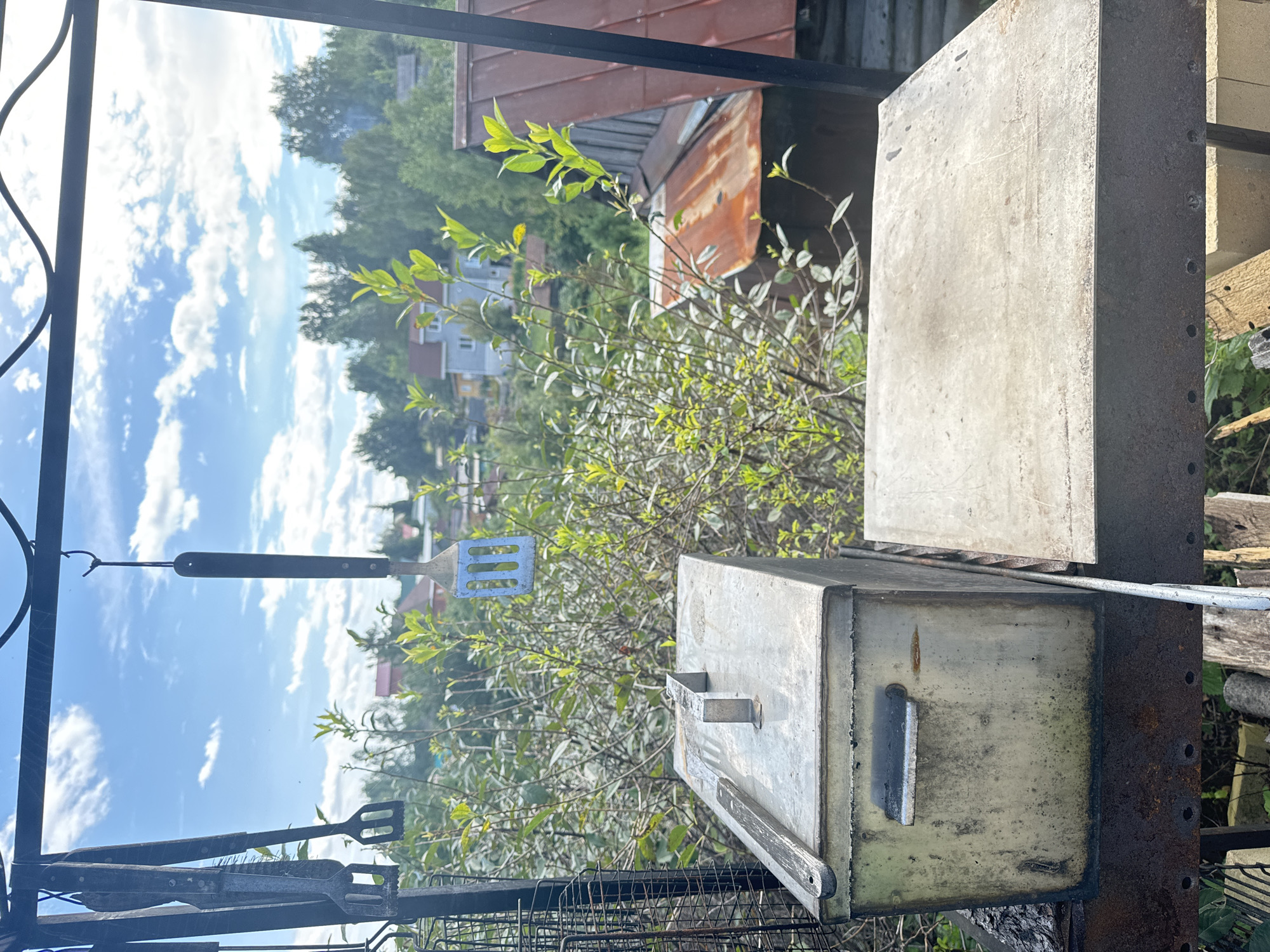 An outdoor scene captures a rustic barbecue setup under a bright, cloudy sky. In the foreground, a weathered metal barbecue box sits on a rusted grate, with a flat metal surface to its right. Above, a metal rack suspends various barbecue tools, including tongs and a spatula. Behind the barbecue equipment, lush green foliage and trees frame a view of houses and rooftops, suggesting a residential area. The overall atmosphere is one of outdoor leisure and a summertime gathering.