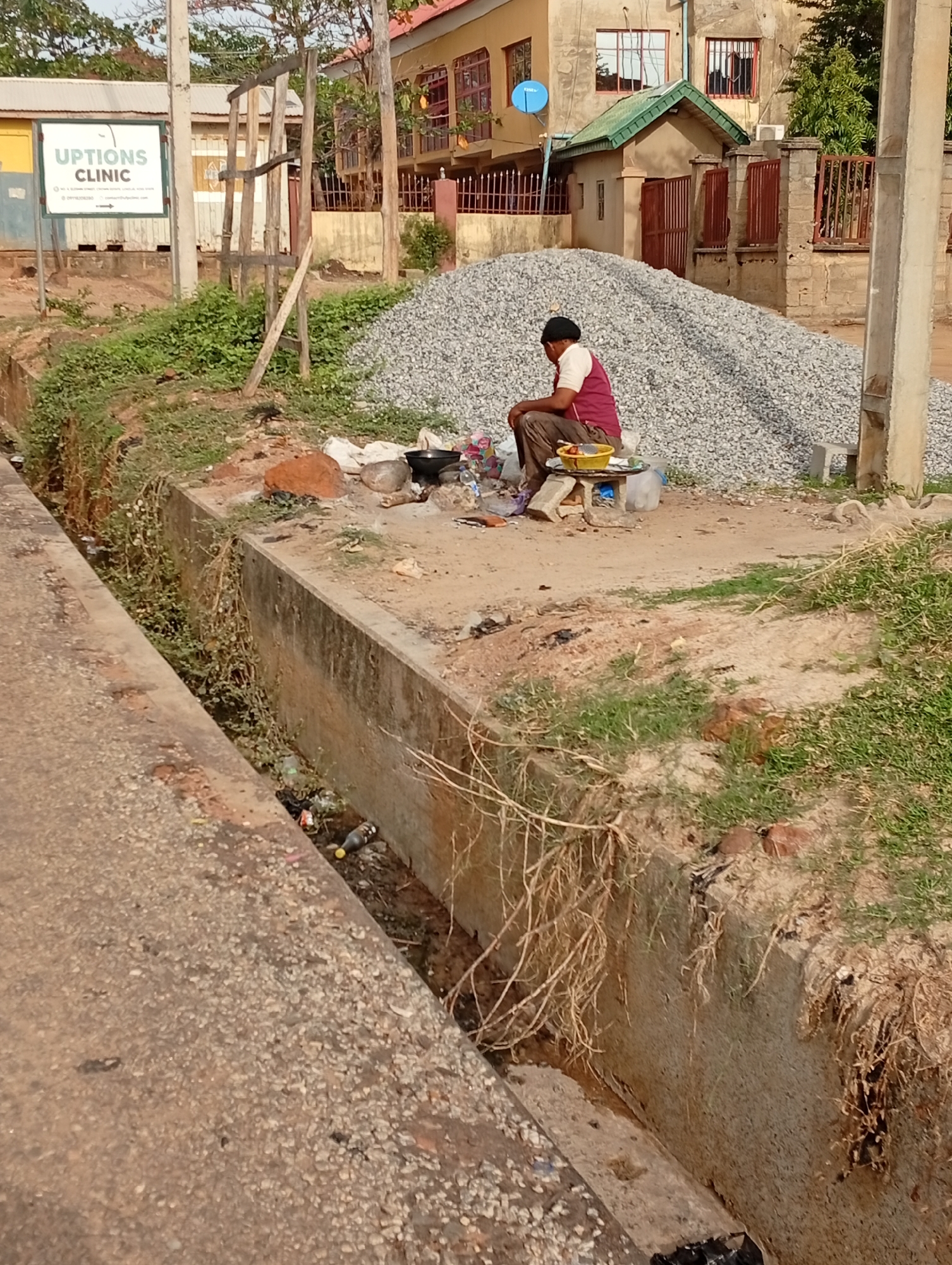 In Adankolo, Nigeria, a man sits on a stool near a pile of gravel, positioned between a concrete drainage ditch and a building. The ditch contains trash and sparse vegetation. Cooking implements are visible near the man. A sign for "UPTIONS CLINIC" is visible in the background. The buildings are a mix of one and two-story structures, some with fences.