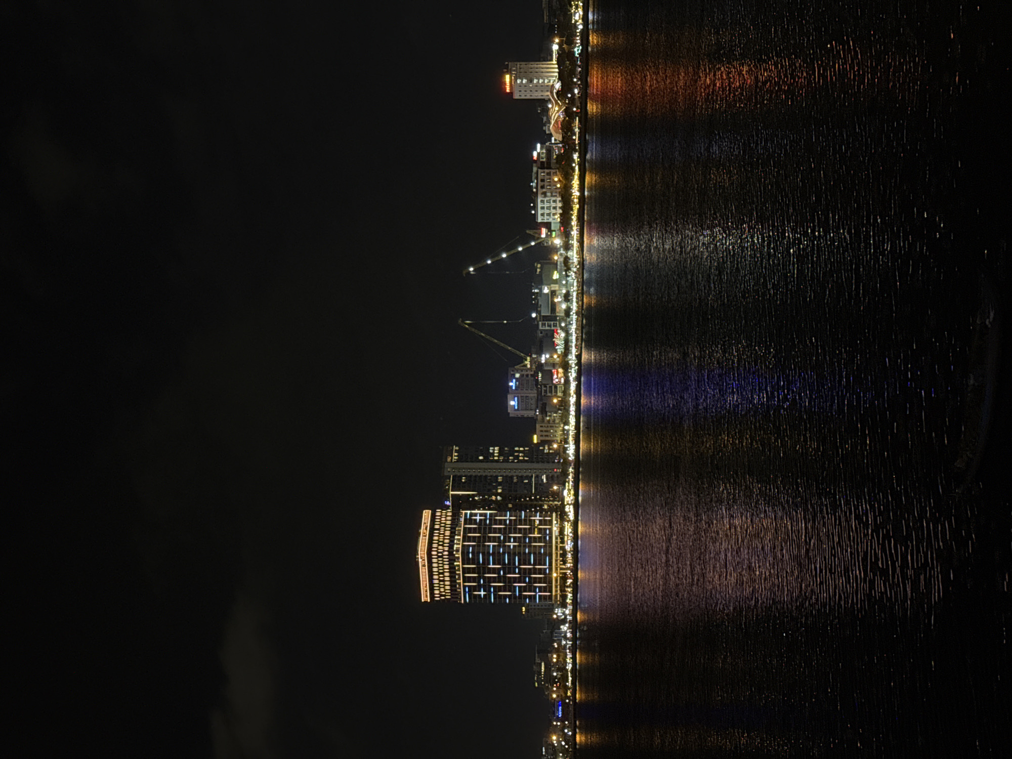 This is a nighttime photograph of the Han River in Da Nang, Vietnam, with the city skyline visible on the left bank. The central subject is a tall, modern building illuminated with a grid of blue and white lights, topped with a band of orange lights. To the right of this building are other structures, including cranes, suggesting ongoing construction or port activity. The city lights are reflected in the dark, rippling water of the river, creating streaks of color. The sky above is dark with a few faint clouds. The overall scene conveys a vibrant, urban nighttime atmosphere.