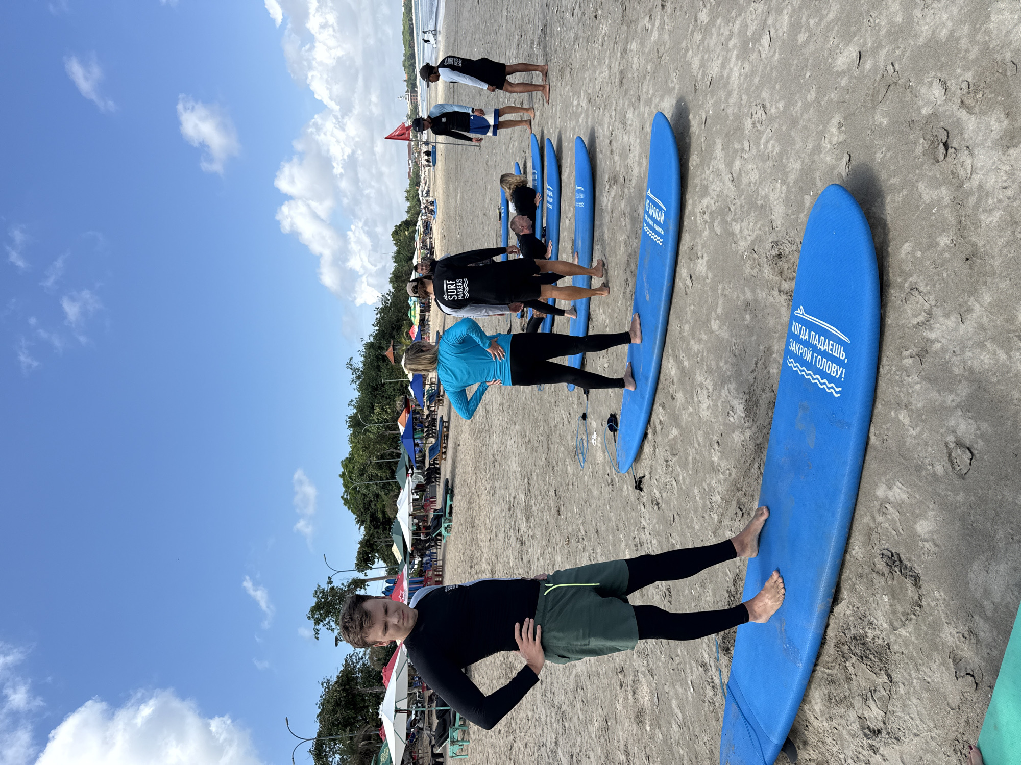On a beach in Kuta, Indonesia, a line of individuals are gathered for what appears to be a surfing lesson. They are standing on or near blue surfboards that are lined up on the sand. The people appear to be a mix of ages and are dressed in swimwear or casual beach attire. The sky is blue with some clouds, suggesting a sunny day. In the background, there are trees and structures, possibly cafes or shops typical of a beach resort area. The overall scene suggests a tourist activity or a surf school session on a popular beach in Kuta.