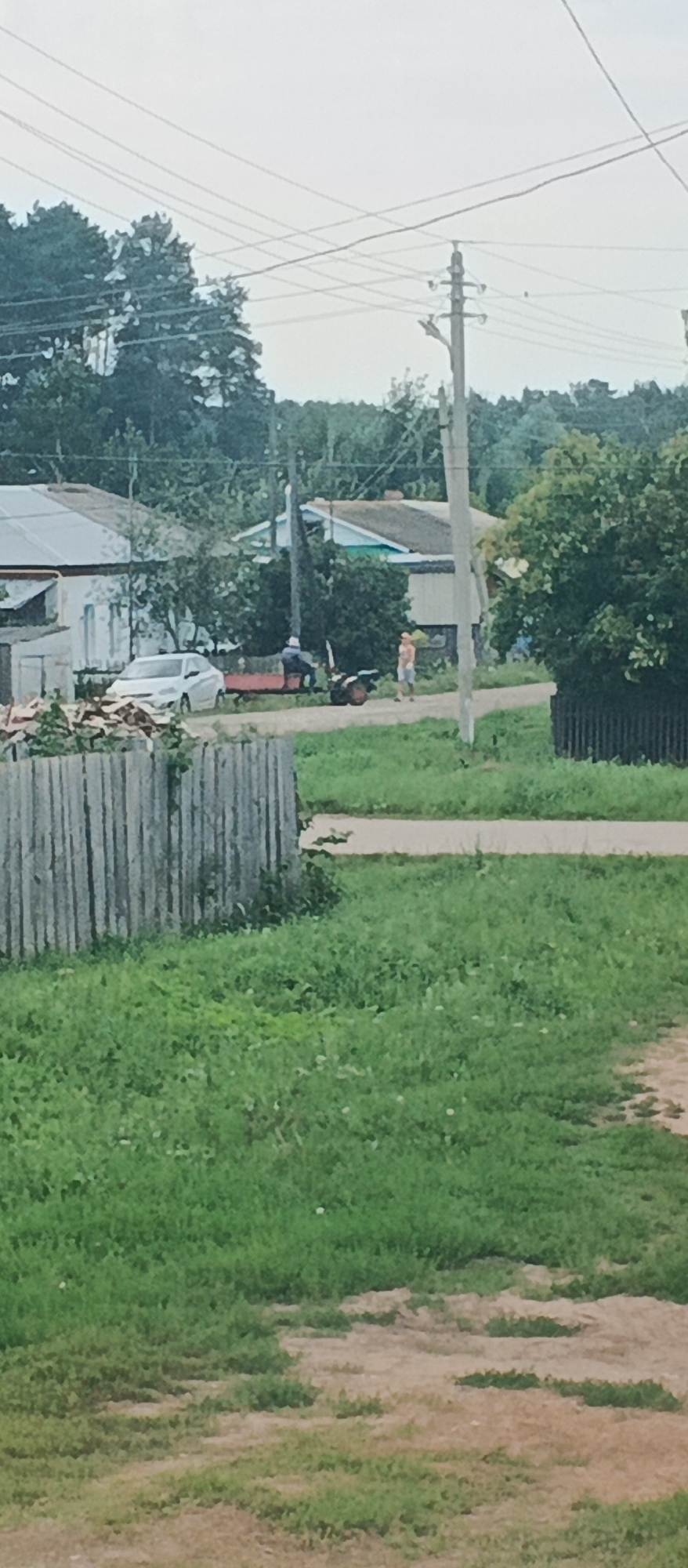 The image depicts a rural scene in the village of Podgornoye, Russia. A white car is parked on a dirt road next to a red trailer. Two individuals are present; one is sitting on the trailer, and the other is standing near a motorcycle. Houses with traditional roofs and fences are visible in the background, along with a power pole and overhead wires. The foreground is dominated by a wooden fence and grassy areas. The sky is overcast.