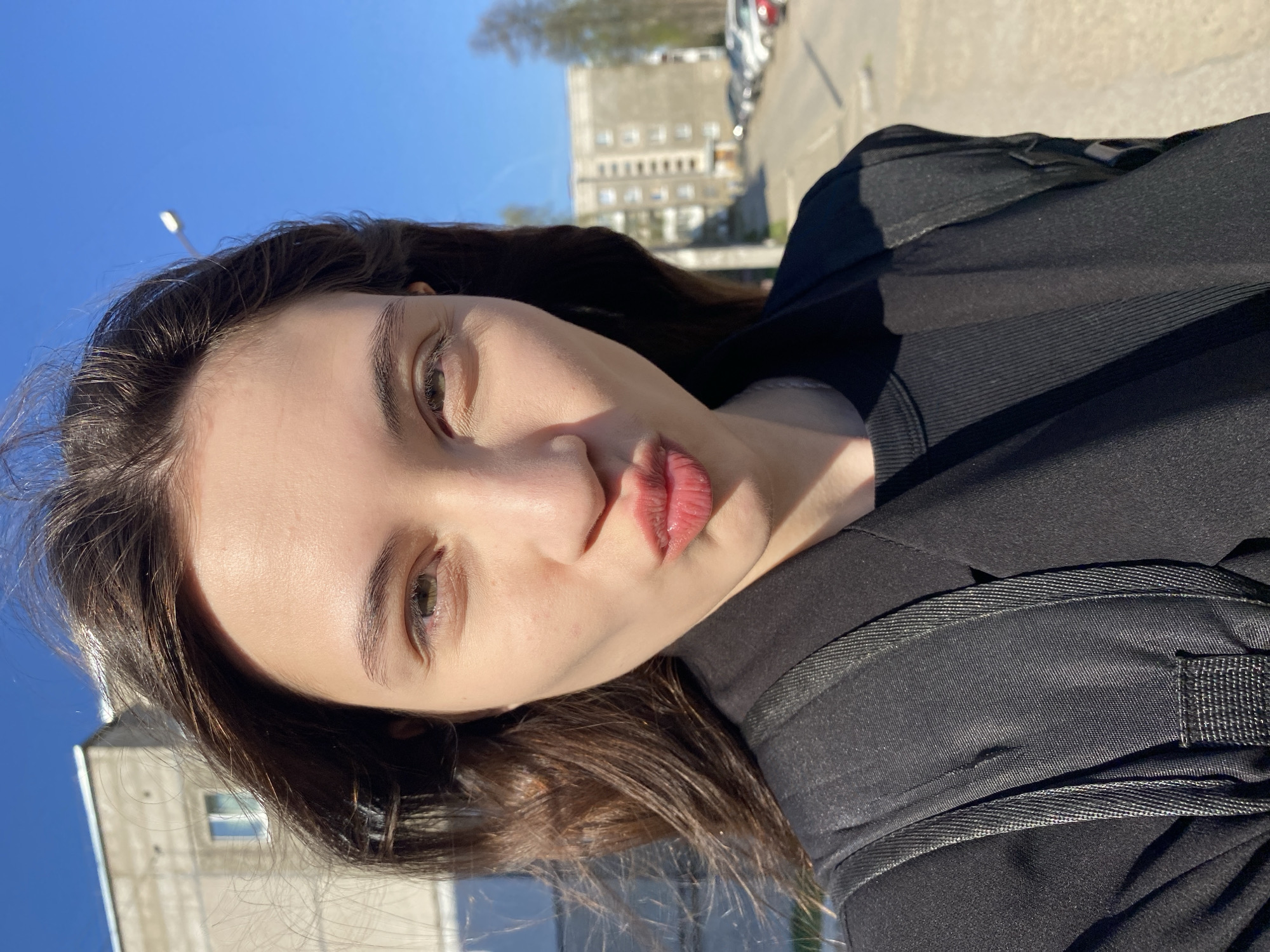 A woman is taking a selfie outdoors, likely in Votkinsk, Russia. She is wearing a black top and backpack. The background shows a clear blue sky, a street with parked cars, and a multi-story building, a common feature of Russian urban environments. The presence of a lamppost further indicates an urban or suburban setting.