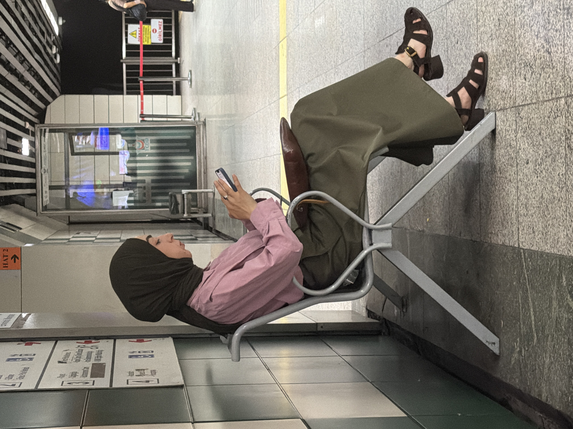 A woman wearing a dark green hijab, a pink collared shirt, and olive green wide-leg pants sits on a metal bench in a metro station. She is looking down at her phone, which she holds in her hands. A brown purse rests on her lap. Her dark brown sandals are visible on her feet. The station appears to be clean with tiled walls and a patterned ceiling. A yellow line is painted on the floor behind the bench. In the background, there is a glass booth with signage that reads "TCDD," indicating it is part of the Turkish State Railways. The overall environment suggests an indoor public transportation hub in Istanbul, Turkey.
