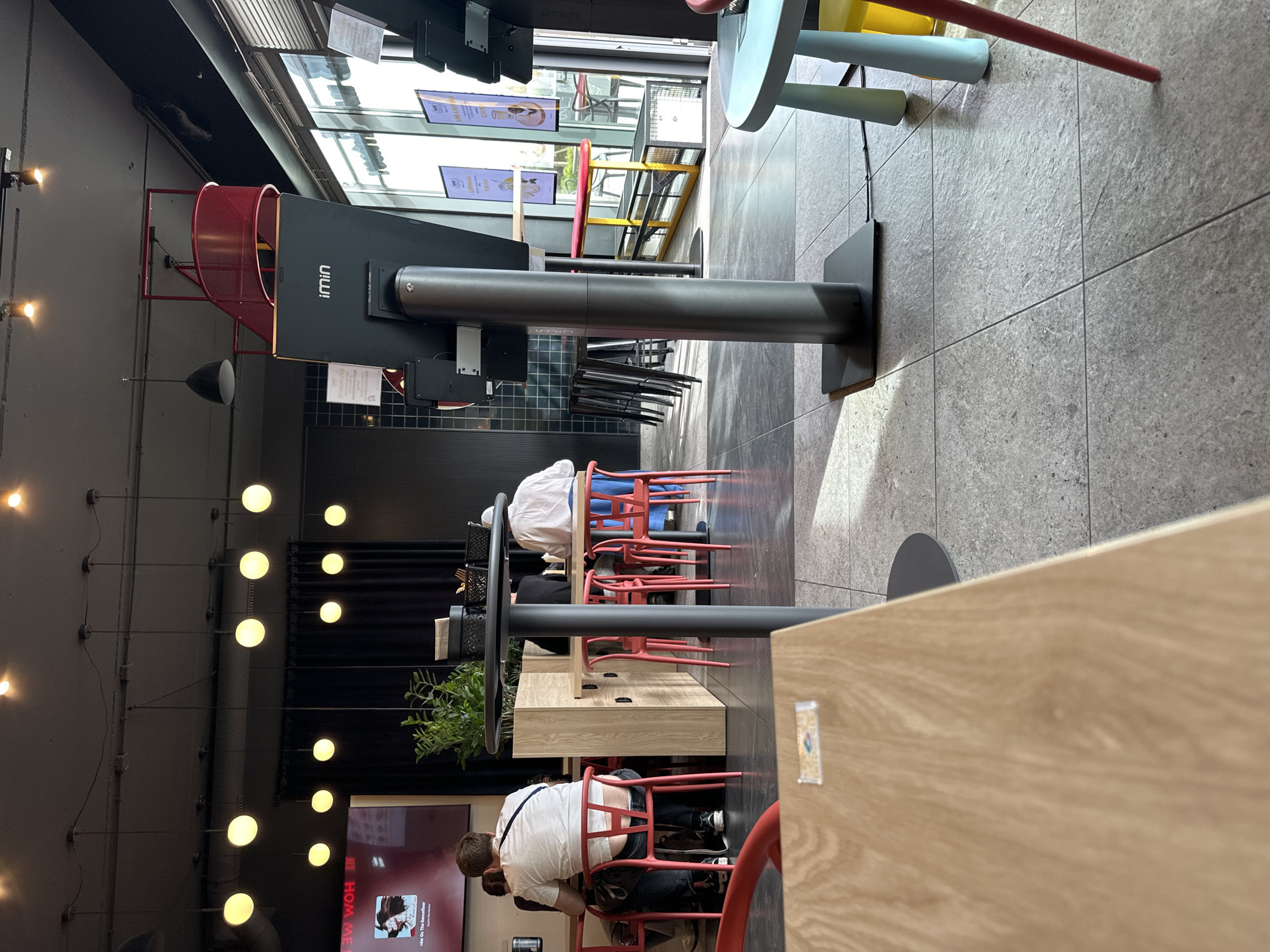 The image depicts the interior of a modern commercial space, likely a cafe or co-working area in Wrocław, Poland. Several individuals are seated at tables, focused on their activities, presumed to be working or socializing.
The environment includes a mix of seating arrangements, with high-top tables featuring colorful stools and more conventional tables with red chairs. Architectural elements include a dark ceiling with exposed lighting fixtures. A black structural component with the "IMIN" logo is present. A television screen with images is visible.
The overall scene suggests a contemporary, functional space designed for work and leisure.