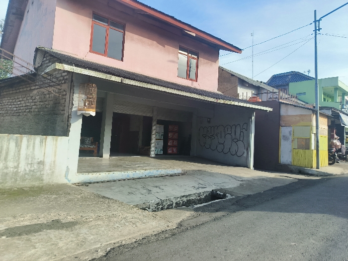The image shows a street scene in Cikalong, Indonesia (latitude: -6.734, longitude: 107.44). In the foreground, a paved street with a drainage ditch runs from left to right. There is a small, shallow ditch in the street. The street is bordered by a concrete sidewalk on the left. A pink two-story building with a pitched roof is on the left side of the image. The building has a small, open-air porch with a concrete floor. There is a birdcage hanging on the side of the building, and a couch is visible in the porch. The side of the building is made of brick and has some white paint on it. The front of the building is painted pink. There are two windows visible on the upper floor of the building. The building has a concrete awning. There is a large, open-air garage area in front of the building. On the right side of the image, there are two buildings with yellow walls and a roof. The buildings have a wooden panel design. There is a small, enclosed area next to the yellow buildings, and a motorcycle is parked in the enclosure. The building on the right has a light green upper floor and is partly obscured by a taller building behind it. The buildings are located on a street in Cikalong, Indonesia. There are electrical wires and a pole in the background. The sky is blue and there are no clouds.