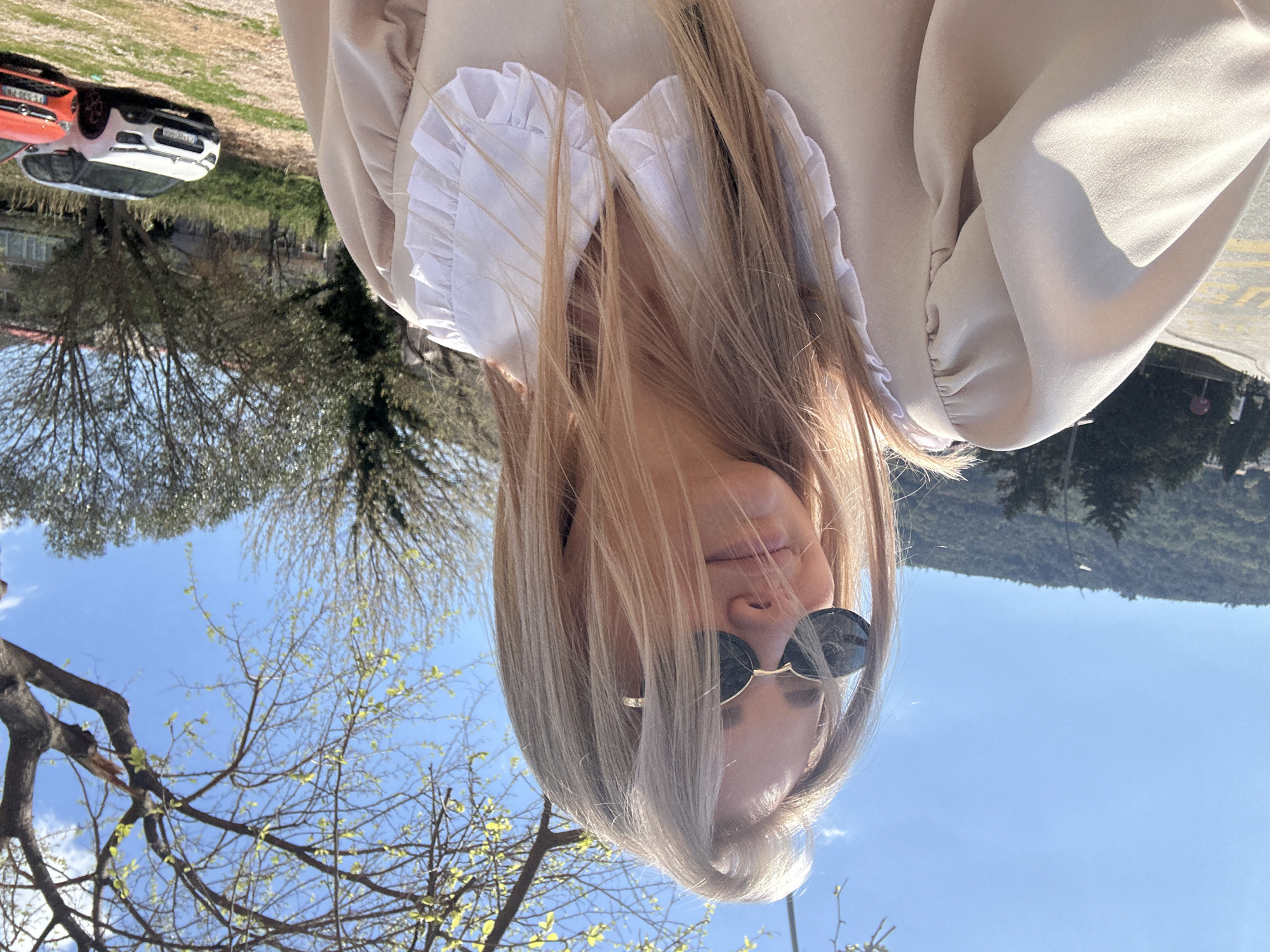 A woman with light brown hair and sunglasses is the focus of the photo, which is taken outdoors in daylight. She is wearing a light-colored dress or top with a ruffled collar. Behind her, the background shows a partly clear blue sky, trees, and a hillside with a mountain in the distance. There are also buildings and cars visible in the background, suggesting an urban or suburban area. The scene may be in Budva, Montenegro.