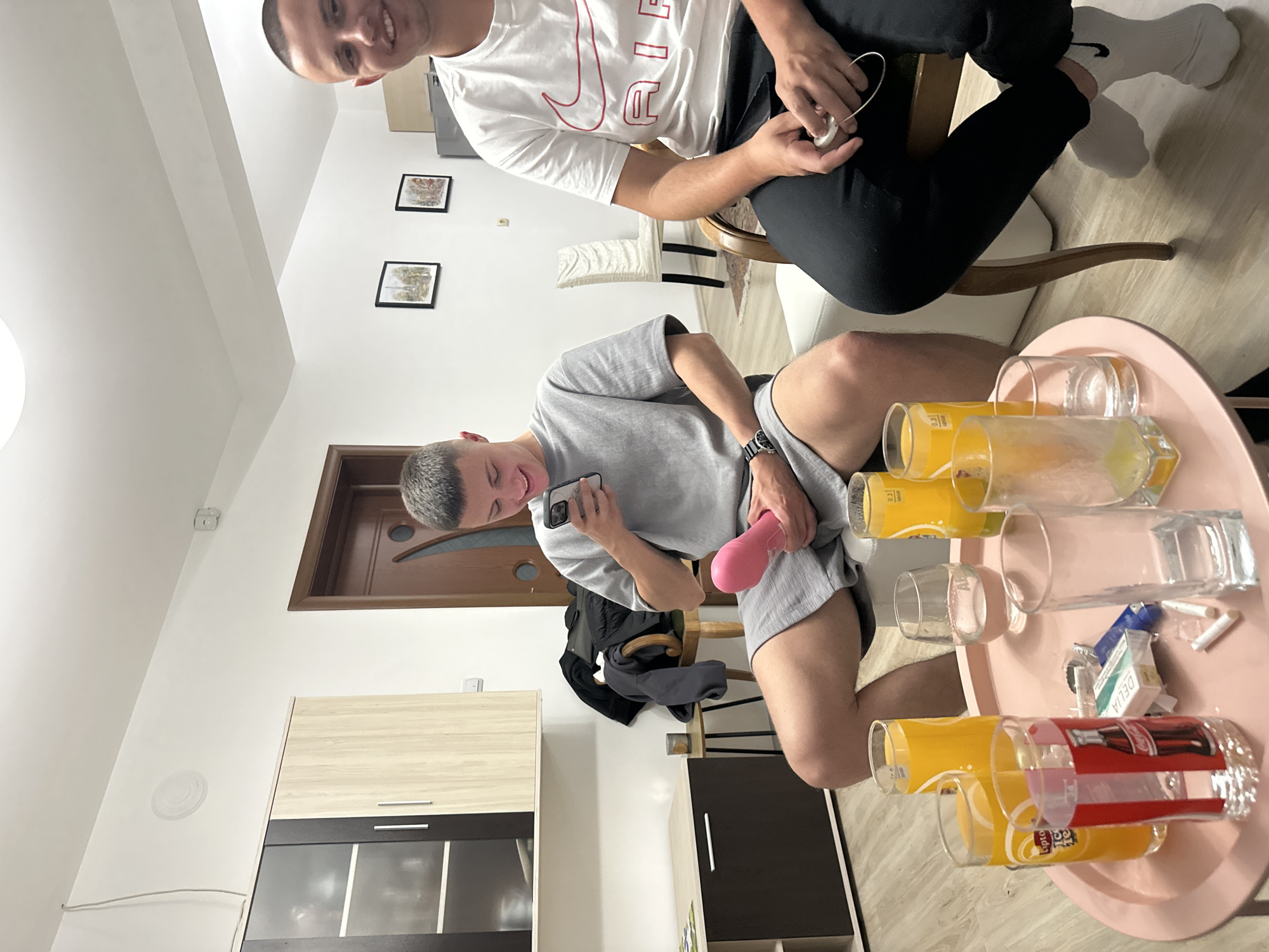 The image depicts two men in an indoor setting. One man is sitting on a chair wearing black pants and a white shirt with a Nike logo. He is holding something in his hands. The other man is in the foreground wearing a grey t-shirt and shorts, looking at a mobile phone and holding a pink object. A round table with glasses and cigarettes is also in the foreground. The location appears to be inside a home or apartment. The city of Graf Ignatievo, Bulgaria is not identifiable from the image.