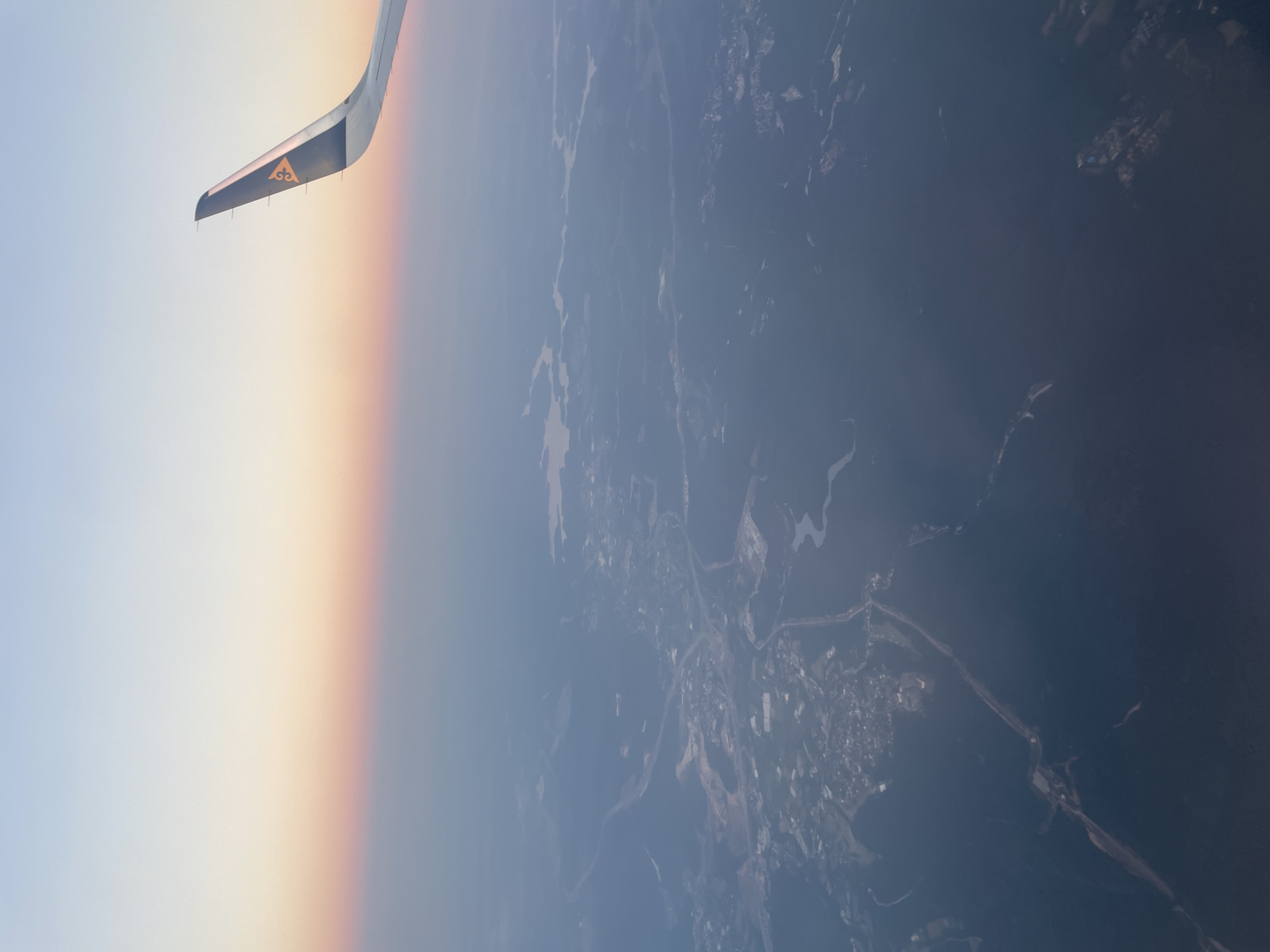 An aerial view shows a landscape below, possibly near Arnavutköy, Turkey, during what appears to be either sunrise or sunset, indicated by the soft gradient of light in the sky transitioning from pale blue to pink and orange. The wing of an airplane, bearing a yellow and black emblem, extends into the upper right corner of the frame, suggesting the image was taken from a flight. The land below is a textured mix of dark green forested areas and lighter patches that could represent urban development, agricultural land, or clearings. Water bodies are visible, appearing as irregular, light-colored shapes interspersed within the landscape, likely lakes or rivers. A network of roads or pathways can be discerned, connecting various developed areas. The overall impression is of a vast, sparsely populated or semi-rural area viewed from a significant altitude.