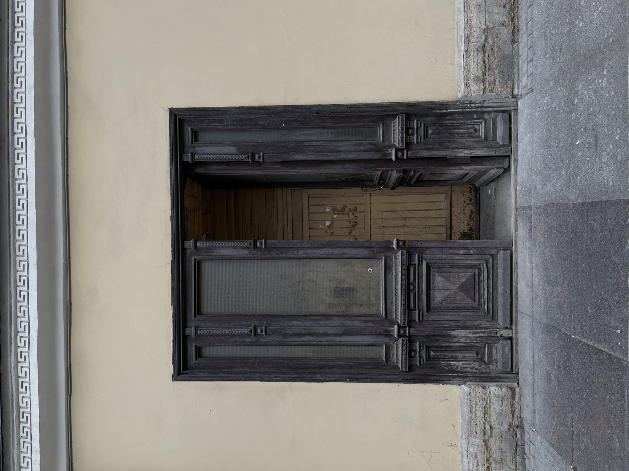 A doorway in Saint Petersburg, Russia, stands ajar, revealing a second, inner door. The exterior door is constructed of dark-stained wood and features decorative molding and panes of glass. The wall surrounding the doorway is painted a light yellow, and a decorative Greek key pattern is visible above the door frame. To the right of the doorway, a gray granite sidewalk is visible.