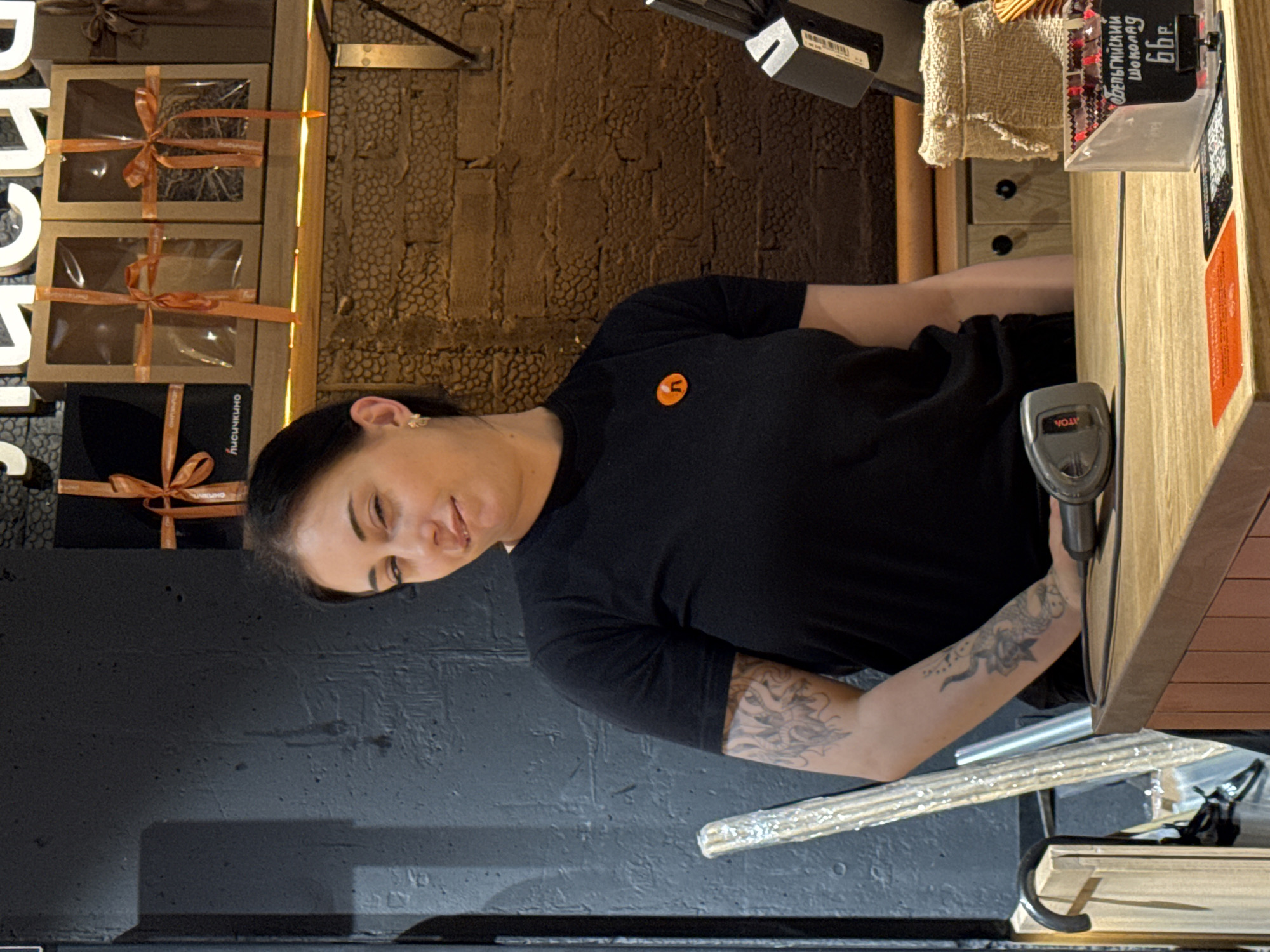 A woman with a tattoo on her arm, wearing a black t-shirt with a small orange logo, is standing in front of a counter. She is holding a scanner. Behind her is a dark gray wall. To her left, boxes with orange ribbons can be seen, and on the right is the counter with product displays. The location is Rostov-on-Don, Russia.
