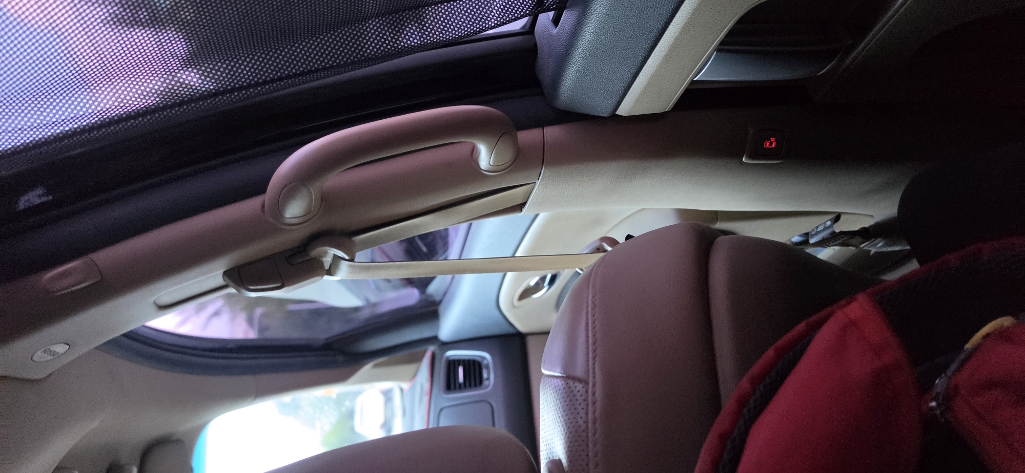 An interior view of a car's passenger side, likely parked in Cheongju-si, South Korea, shows a beige grab handle attached to the car's pillar. A beige seatbelt is visible hanging down from the handle. A brown leather car seat is partially visible on the left side of the image. A red backpack with black trim rests on the floor in the lower left corner. The window on the right side of the car is covered by a black mesh sunshade, revealing a blurry, purplish exterior light. A small red indicator light, possibly a door lock symbol, is illuminated on the lower part of the pillar. The scene suggests a moment of stillness within the vehicle, possibly during a stop.