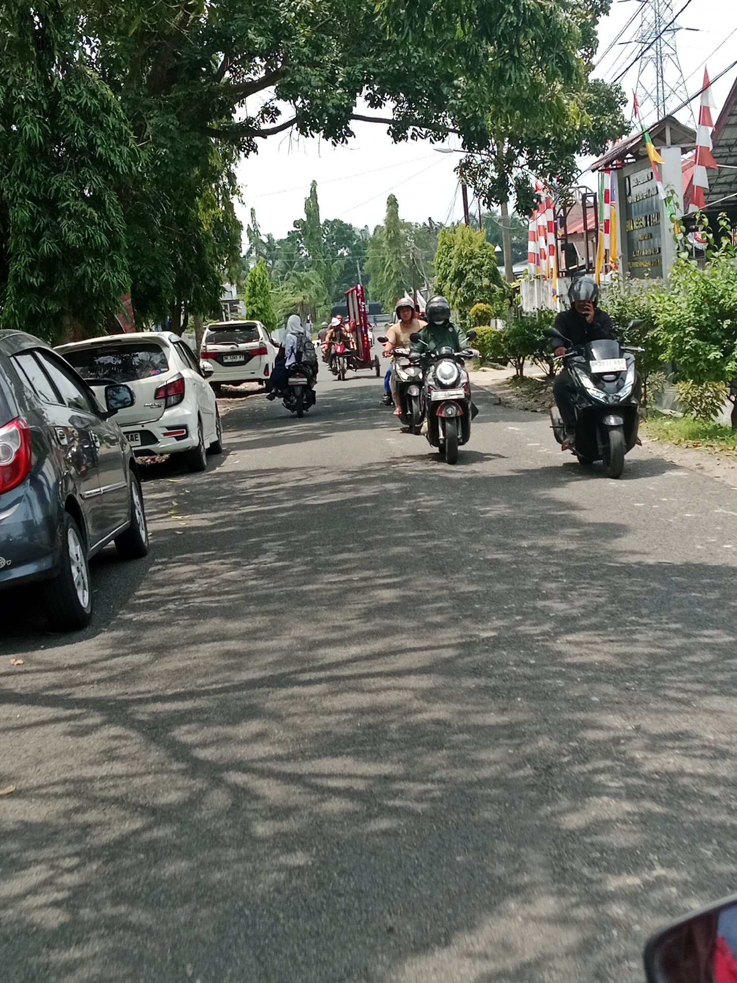 The image depicts a street scene in the City of Binjai, Indonesia, on a sunny day with dappled sunlight filtering through trees onto the asphalt road. Several motorcycles are in motion, with riders wearing helmets. In the foreground, a dark-colored car is parked on the left side of the road, and a white car is parked further down. On the right side of the road, there are buildings with flags and signage, suggesting a commercial or civic area. Tall trees line the left side of the street, and green bushes and trees are visible on the right. The overall impression is that of a typical urban street with ongoing traffic and activity.