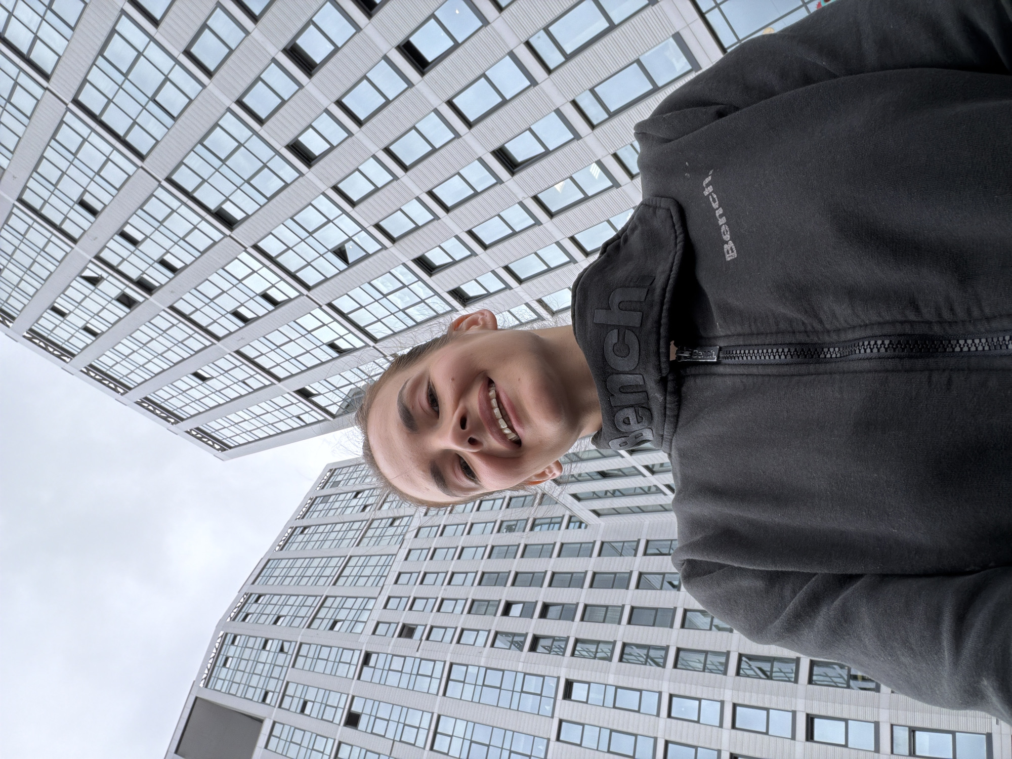 A low-angle, medium shot captures a woman with a wide smile and her teeth visible, looking directly into the camera. She is wearing a black zip-up sweatshirt with the brand "Bench" visible on the chest and collar. The background features the facades of two modern, high-rise buildings with numerous windows, set against an overcast sky. The architecture suggests an urban setting. The image is a selfie, taken by the subject. The location is identified as Ufa, Russia.
