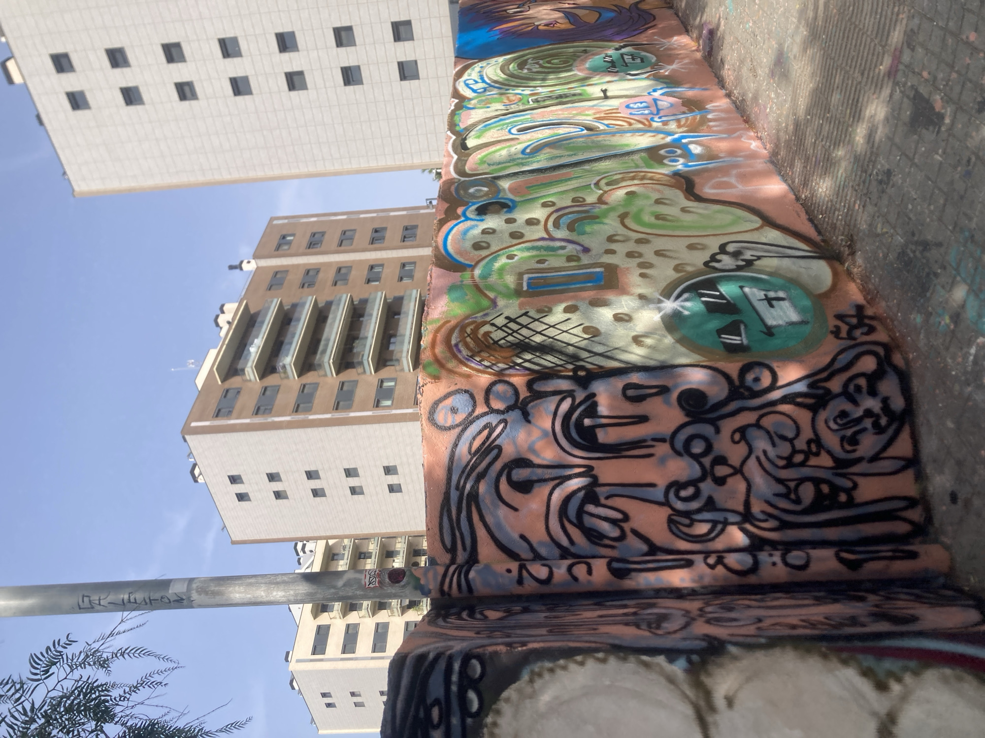 The image captures a section of a graffiti-covered wall in Barcelona, Spain. The wall is painted with various colorful and abstract designs, including cartoonish faces and lettering. In the background, modern apartment buildings rise against a clear blue sky. A lamppost stands near the wall, also bearing some graffiti. The ground is paved with cobblestones, and dappled sunlight casts shadows on the scene, indicating an outdoor urban environment. The overall impression is one of urban art and architecture.