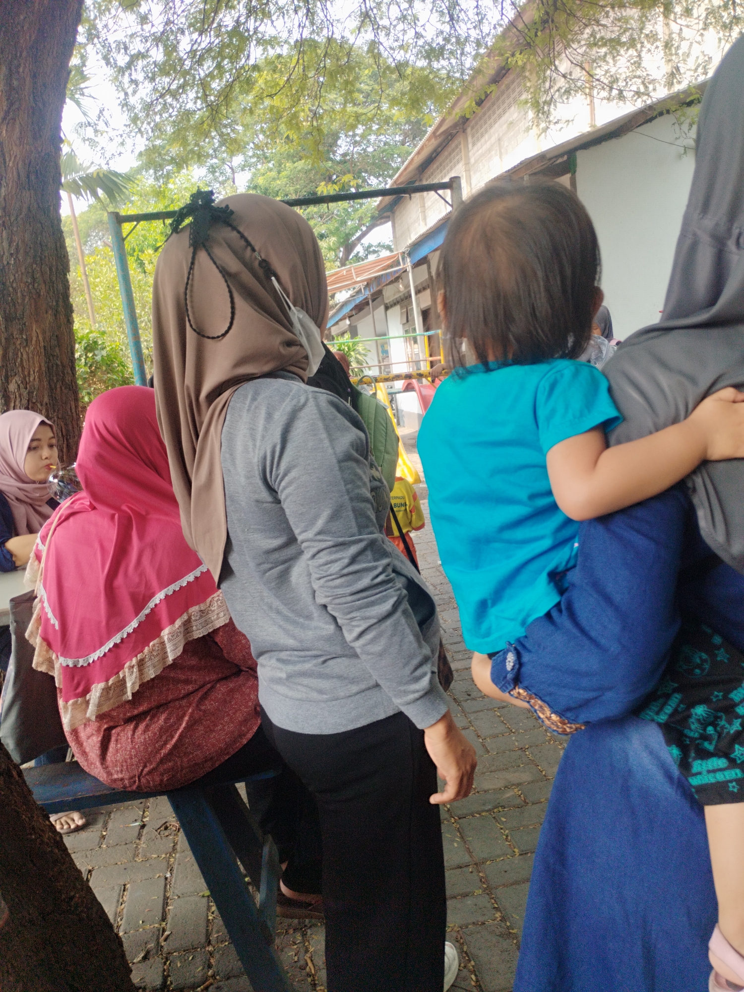 The image depicts an outdoor scene, likely a public space in Surabaya, Indonesia, with several individuals present. In the foreground, a person wearing a brown hijab and a gray long-sleeved shirt is seen from behind, their face obscured. They are holding a child who is wearing a bright blue shirt and dark blue pants, also viewed from the back. To the left, another person in a pink hijab and a patterned skirt is seated on a blue bench. The ground is paved with interlocking bricks, and fallen leaves are scattered around. In the background, there is a building with a light-colored facade and a metal roof, partially obscured by trees with green foliage. A portion of a tree trunk is visible on the far left. The overall impression is of a casual gathering in an urban outdoor setting.
