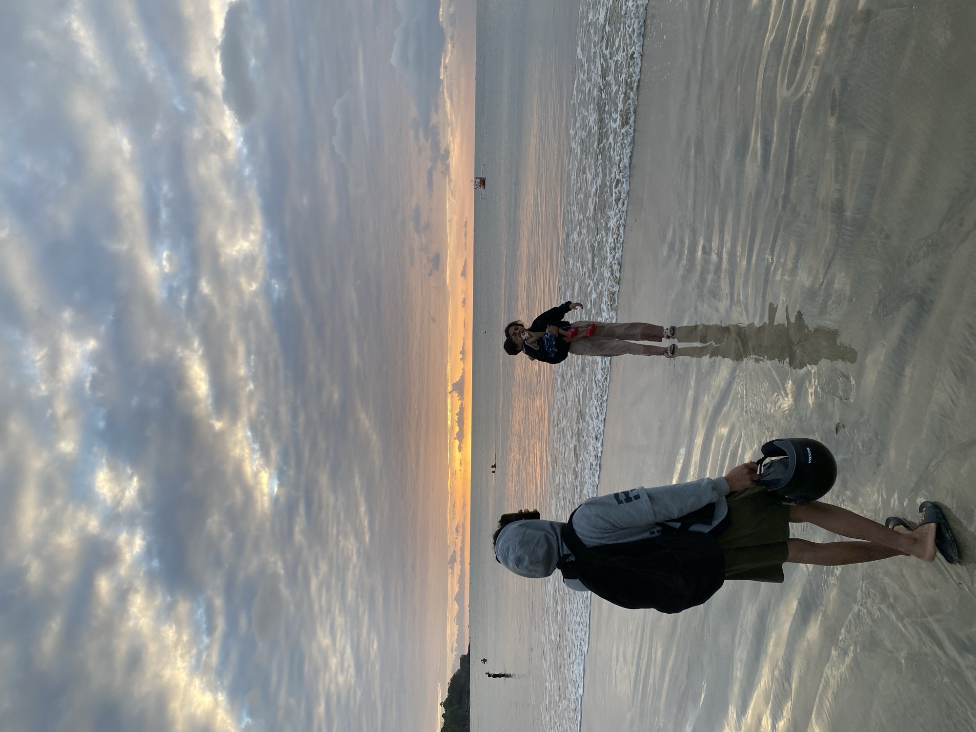 At dusk on the beach in Kuta Selatan, Indonesia, a young person with a backpack and a helmet stands on the wet sand, facing away from the camera. The person wears a grey hooded sweatshirt, olive green shorts, and black flip-flops. Further down the beach, a woman holds a child and is looking out at the ocean. The water has small waves and there are a few boats visible in the distance. The sky is filled with dramatic clouds, illuminated by the setting sun, casting a warm glow over the scene. The wet sand reflects the colors of the sky, creating a shimmering surface.