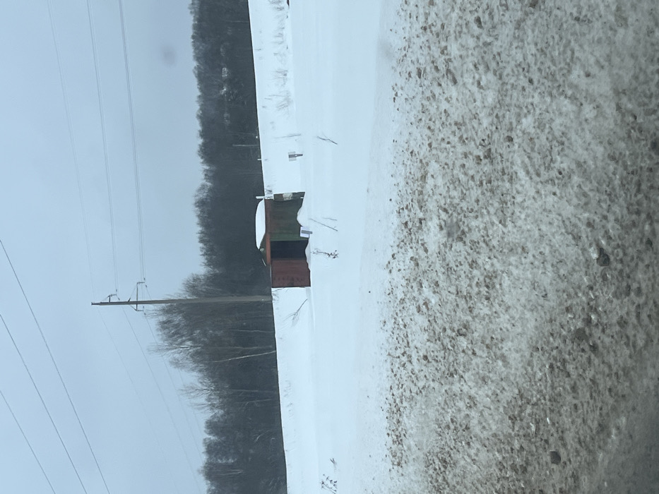 This image depicts a snow-covered rural landscape in the city of Poddubki, Russia, under an overcast gray sky. The foreground presents a textured expanse of dirty snow intermingled with brown gravel and dirt, indicating a disturbed ground area. The midground is dominated by an unbroken field of white snow, from which several thin, leafless plant stems protrude. Centrally positioned in this snowfield is a small, rectangular, shed-like structure, partially embedded in the snow. Its visible left panel is a rusted reddish-brown, while the right panel is dark green, and its flat roof is topped with snow. A dark, open rectangular entrance is visible on its front face. Two small, white rectangular markers are partially visible in the snow directly in front of the structure, and a darker, square-topped marker on a short pole stands further to the right in the snow. The background features a dense, dark line of leafless deciduous trees. Above the tree line, a tall utility pole with multiple horizontal crossarms supports several power lines against the uniformly gray sky. No individuals, animals, or active subjects are present.