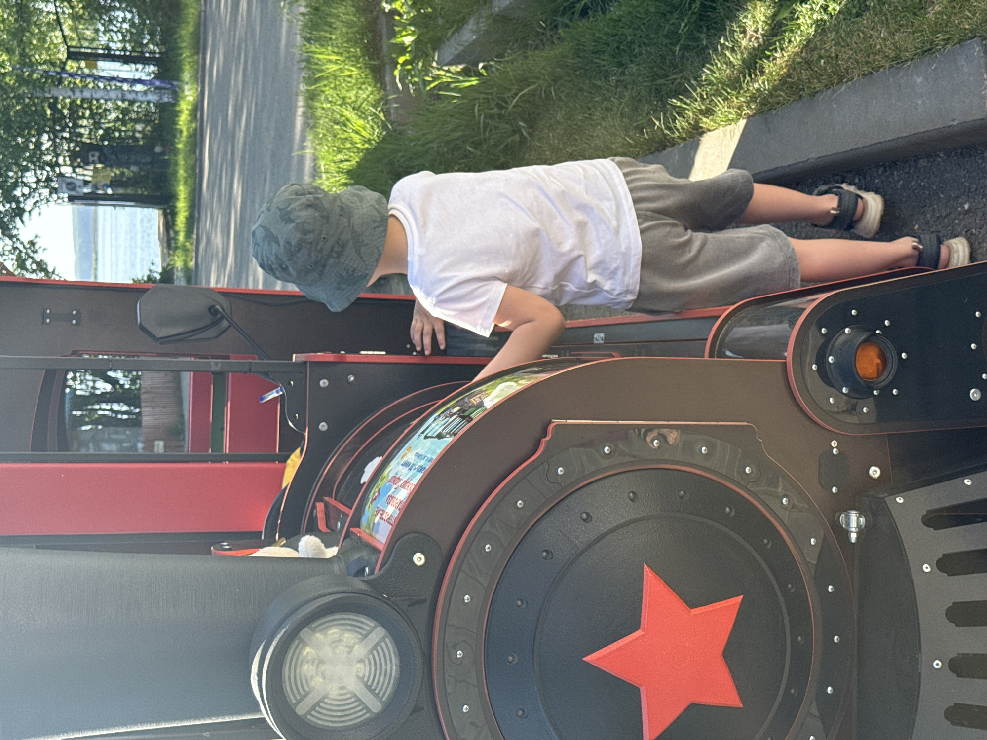 A child wearing a white shirt, gray shorts, and a camouflage bucket hat is lying on a playground train in the city of Chaikovsky, Russia. The train is red and black with a red star on the wheel. The surrounding environment includes grass, a road, and trees. The child is oriented towards the front of the train and appears to be interacting with the controls or details inside the train's cabin.