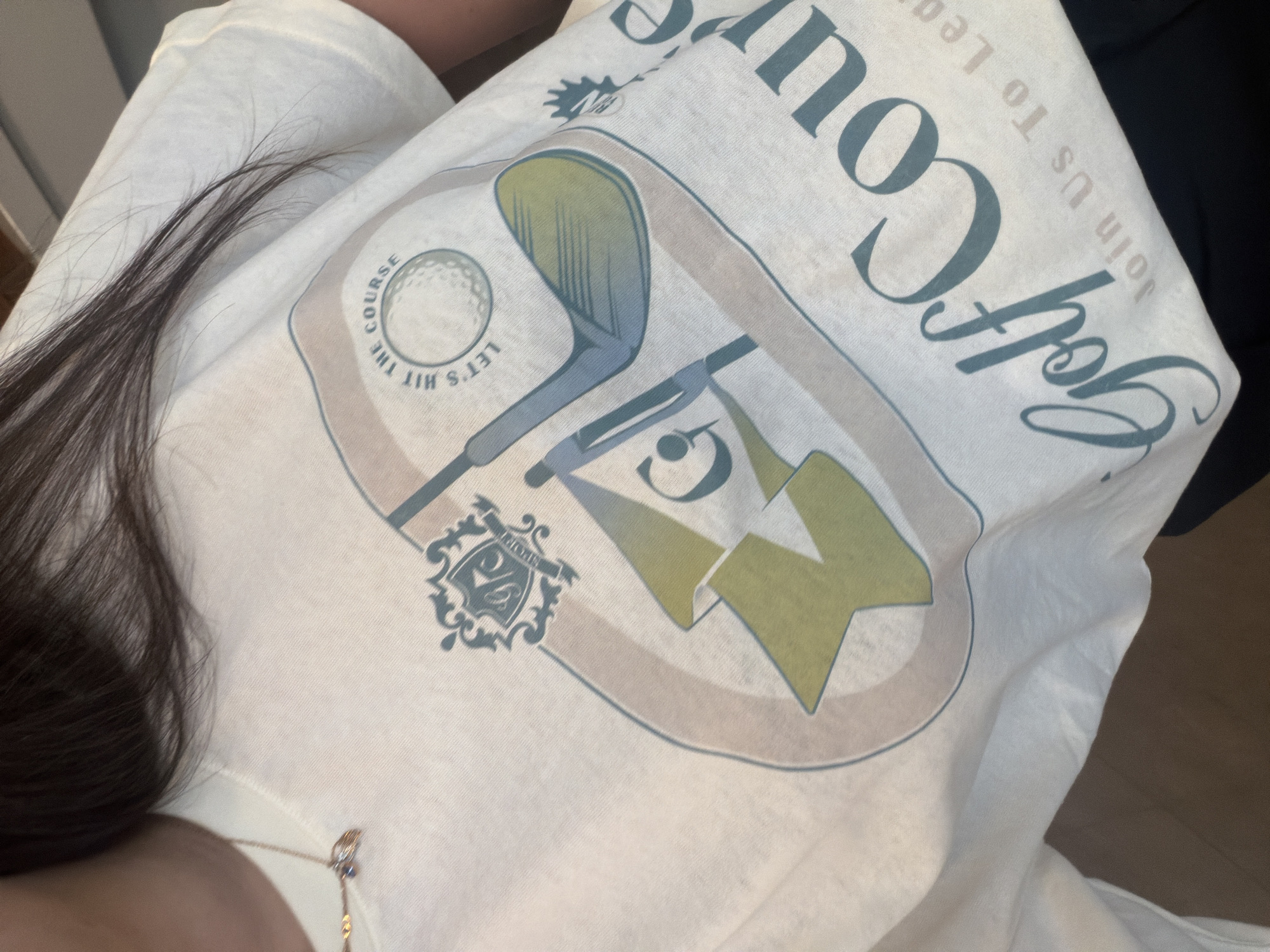 A close-up shot shows a person wearing a white t-shirt with a golf-themed design. The design includes the words "Golf Course Join Us To Lead", a golf club, a golf ball, and green chevron shapes, all enclosed within a badge-like emblem. Part of a necklace is visible near the person's neck. The location is not discernible from the image.