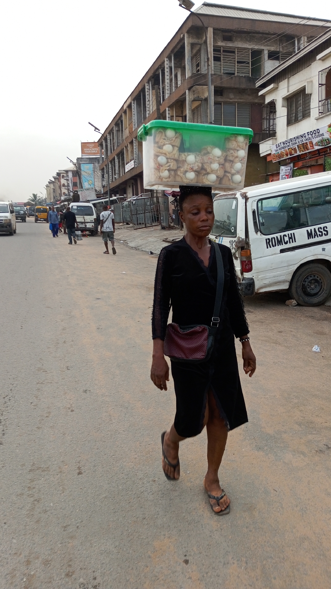 A woman walks along a paved street in Onitsha, Nigeria. She carries a green-lidded plastic container filled with wrapped items topped with white eggs on her head. She wears a black, long-sleeved dress, a small maroon-colored purse on a strap across her body, and dark-colored flip-flops. In the background, a white van with "ROMCHI MASS" written on its side is parked on the right. Other pedestrians and vehicles are visible in the street. Buildings line the street on both sides, with storefronts and signage visible. The sky is overcast.