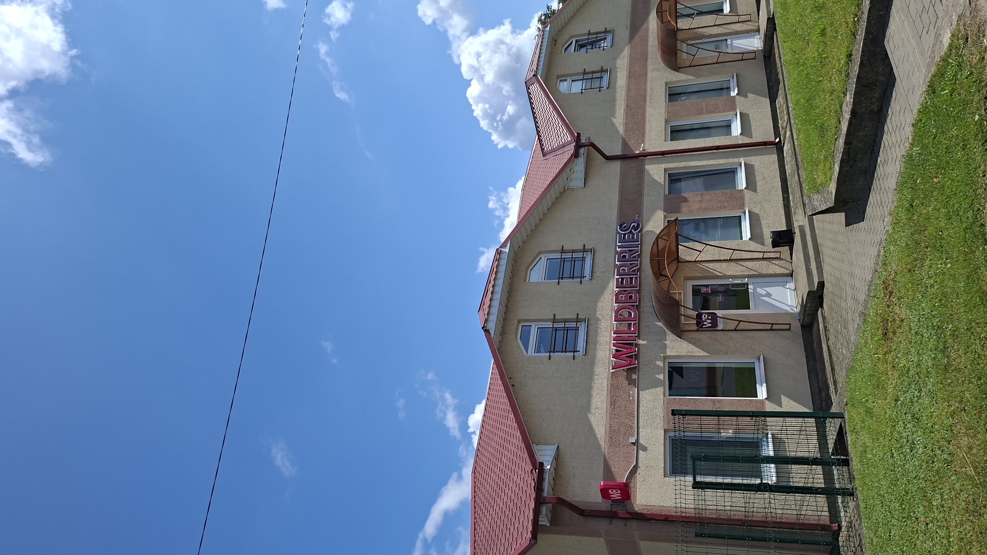 The image depicts the Wildberries store, an e-commerce and retail company, located in the city of Myadzyel, Belarus. The storefront features prominent signage with the "WILDBERRIES" logo in bright pink lettering. The building has a light-colored facade with a red tiled roof and multiple windows. A green metal fence is visible in front of the building, along with a paved walkway and a grassy area. The sky is blue with scattered white clouds.