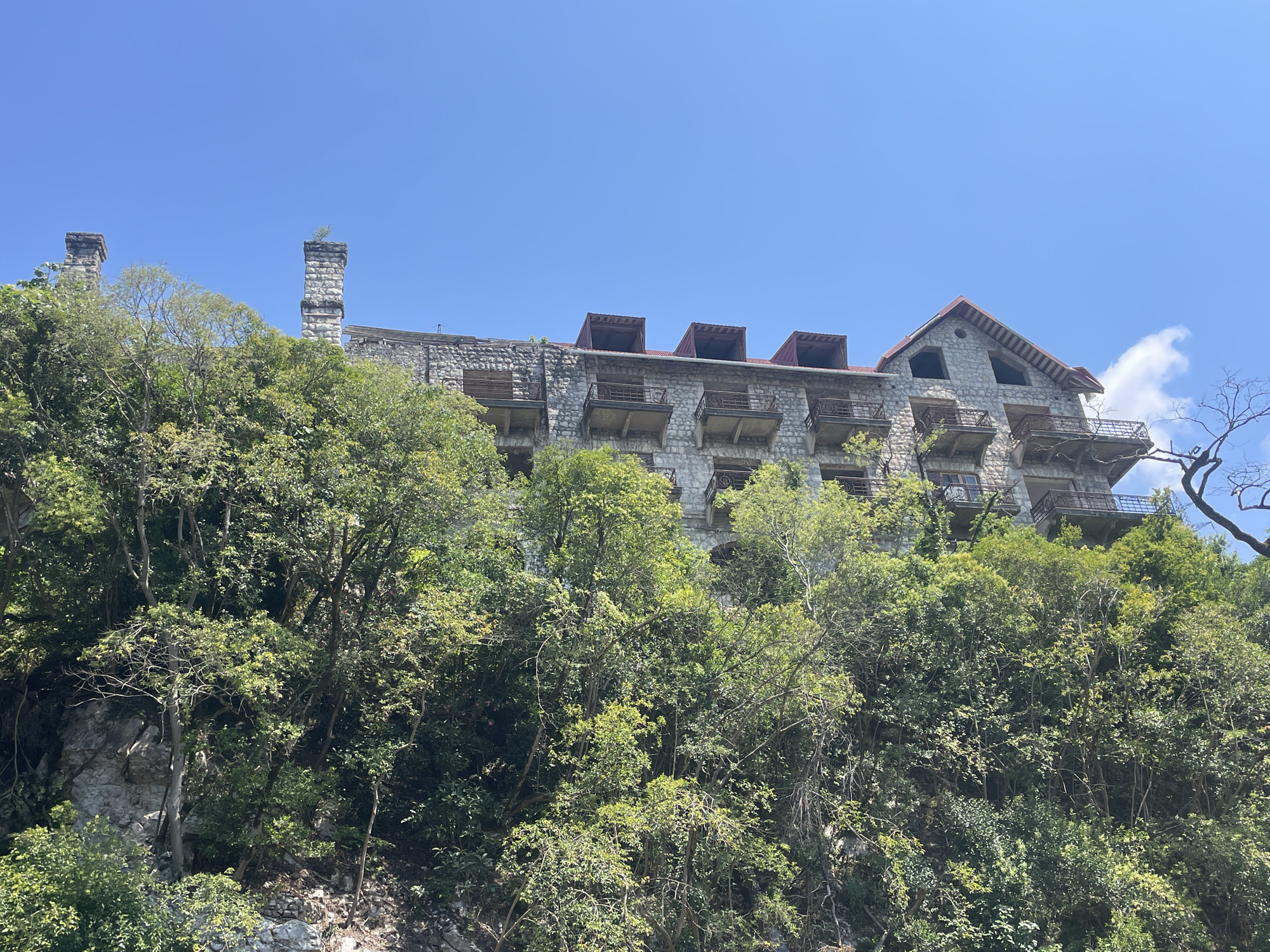 The image depicts an abandoned stone hotel situated on a steep, tree-covered slope. The hotel features multiple stories with balconies and a red, gabled roof with dormer windows. Two stone chimneys rise from the rooftop, one of which has a small plant growing on top. The building appears weathered and in disrepair, with some balconies showing signs of decay and windows boarded up or missing. The surrounding environment is lush with dense green foliage and rocky terrain. The sky above is clear and blue with a few wispy clouds visible on the right side of the image. The scene suggests a remote and perhaps overgrown location, consistent with the description of Abaata, Abkhazia, known for its abandoned structures and natural beauty. There are no people or visible signs of recent activity.