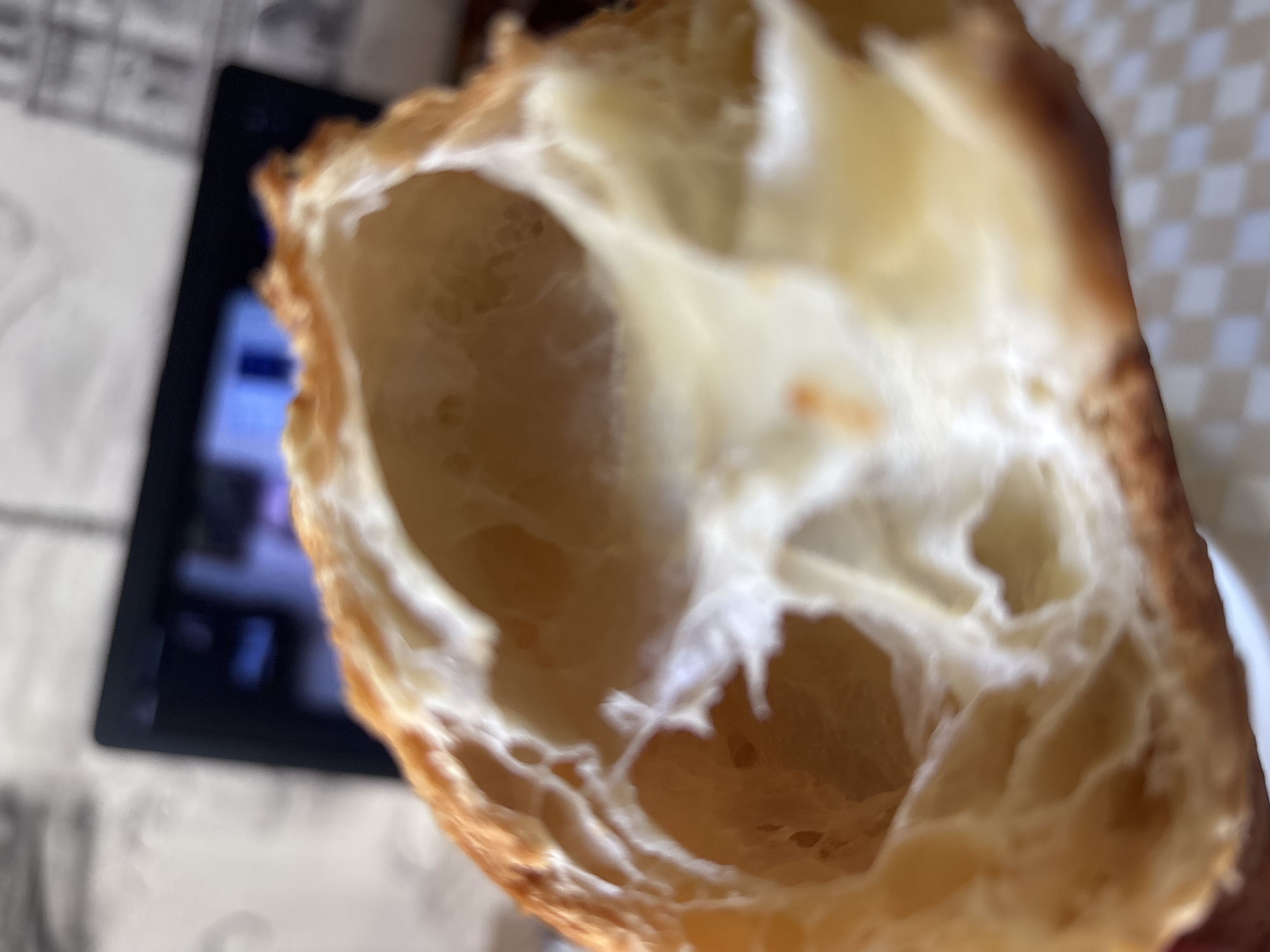 A close-up, shallow depth-of-field shot focuses on the interior of a flaky, golden-brown pastry, likely a croissant. The pastry's layers are visible, with airy pockets and a slightly glossy surface. The background is blurred, showing what appears to be a laptop screen reflecting light and a textured, checkered surface beneath. There is no indication of the city of Bila Tserkva, Ukraine, or any people or specific actions.