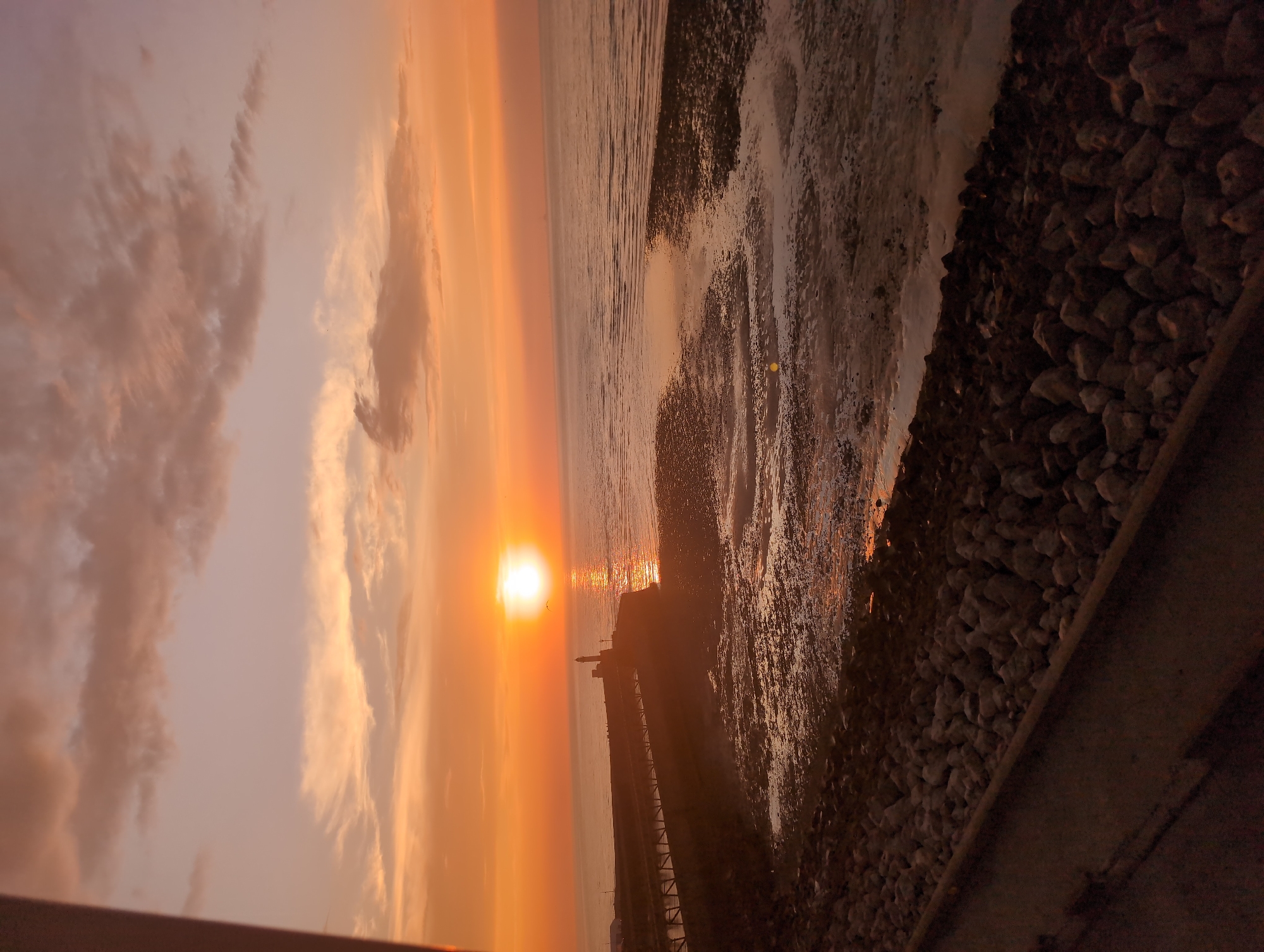 This image depicts a sunset scene at the coast, likely Le Tréport, France, given its distinctive lighthouse. The sun, a bright orb with a golden halo, is low on the horizon, casting a warm, orange glow across the sky and reflecting on the calm sea. Wispy, pinkish clouds dot the sky, adding to the serene atmosphere.
In the mid-ground, a long, narrow pier extends from the land into the sea, culminating in a lighthouse. The pier appears to be constructed of wood and metal, with a railing along its side. The lighthouse, a tall, slender structure, stands prominently at the end of the pier. The foreground features a rocky shoreline, with large, irregular stones forming a natural barrier. The tide appears to be out, revealing a wet, sandy expanse with shallow pools of water. The overall scene is tranquil and picturesque, evoking a sense of peace and natural beauty. There are no people or animals visible in the image. The lighting is soft and diffused, characteristic of a sunset.