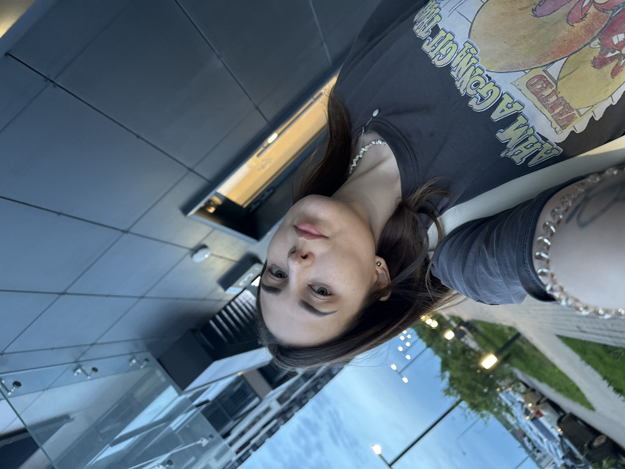 The image is a selfie taken from a low angle, depicting a woman with brown hair and green eyes. She is wearing a black t-shirt with a graphic print, a pearl necklace, and a spiked silver bracelet. The background suggests an urban environment. The architecture visible is modern, featuring a black building with reflective glass panels and lights. Based on visual cues like architectural styles and infrastructure, the location could potentially be Perm, Russia. The presence of green vegetation and streetlights indicates an outdoor setting, possibly a street or plaza.