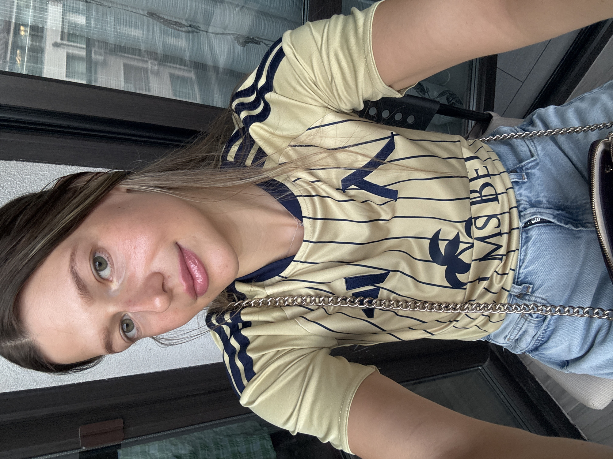 A young woman with blonde hair and green eyes is taking a selfie. She is wearing a gold and navy striped athletic shirt and denim shorts. A silver chain purse is visible. The background shows a window with a city view, possibly from a balcony, suggesting an urban setting in Sofia, Bulgaria.