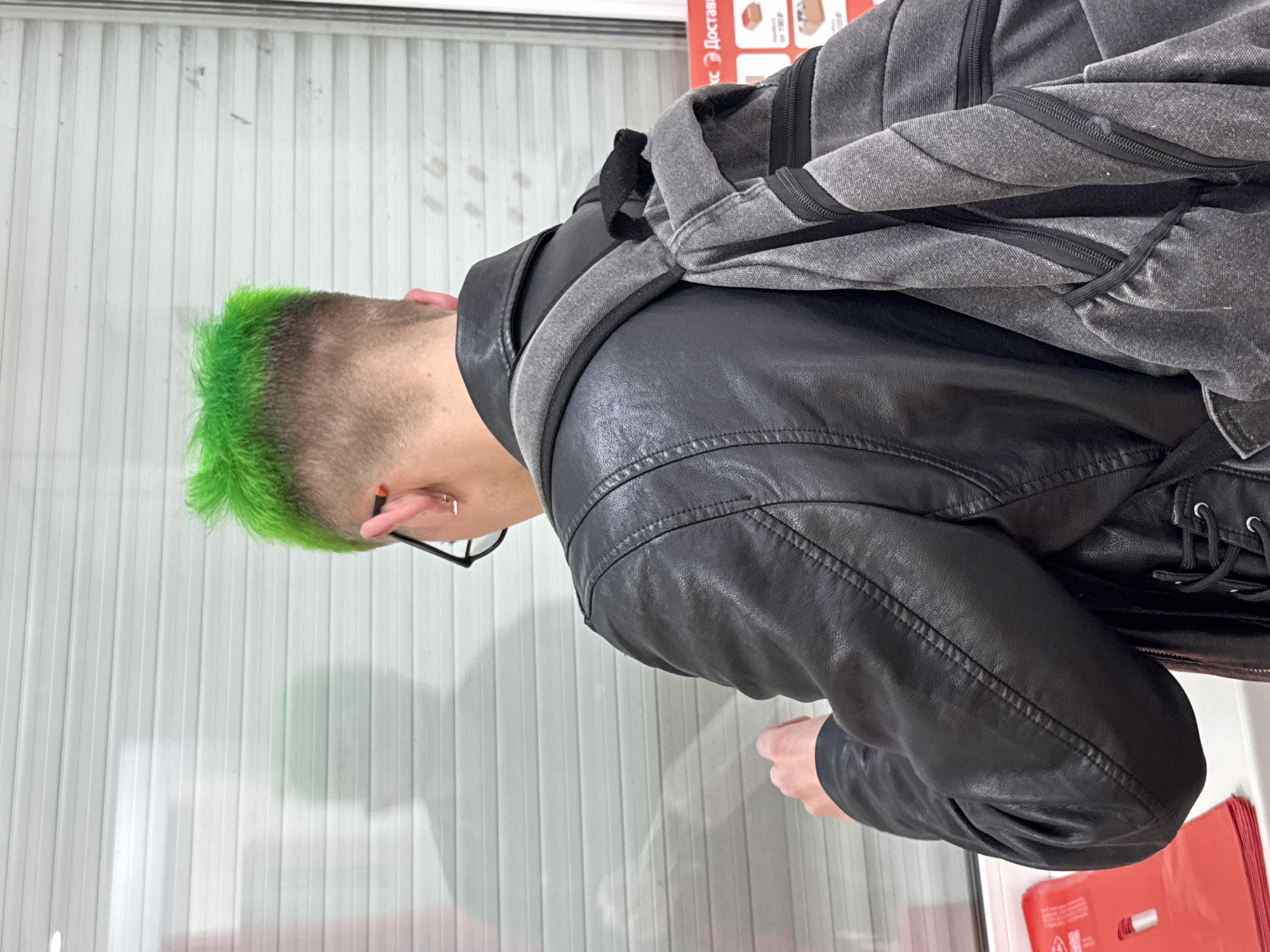 A person with bright green spiked hair is viewed from behind, wearing a black leather jacket and a gray backpack. They are standing in front of a white, ribbed shutter-like surface, possibly a window or door. A shadow of the person is visible on the surface. In the foreground to the left, a stack of red folders or boxes is partially visible. To the right, a blurry sign with Russian text that appears to read "Доставк" (likely part of "Доставка," meaning delivery) and some price tags is visible, suggesting a commercial or service location. The overall environment appears to be indoors, possibly a shop or a transit point. There is no specific indication of the location being in Saint Petersburg, Russia, other than the presence of Russian text.