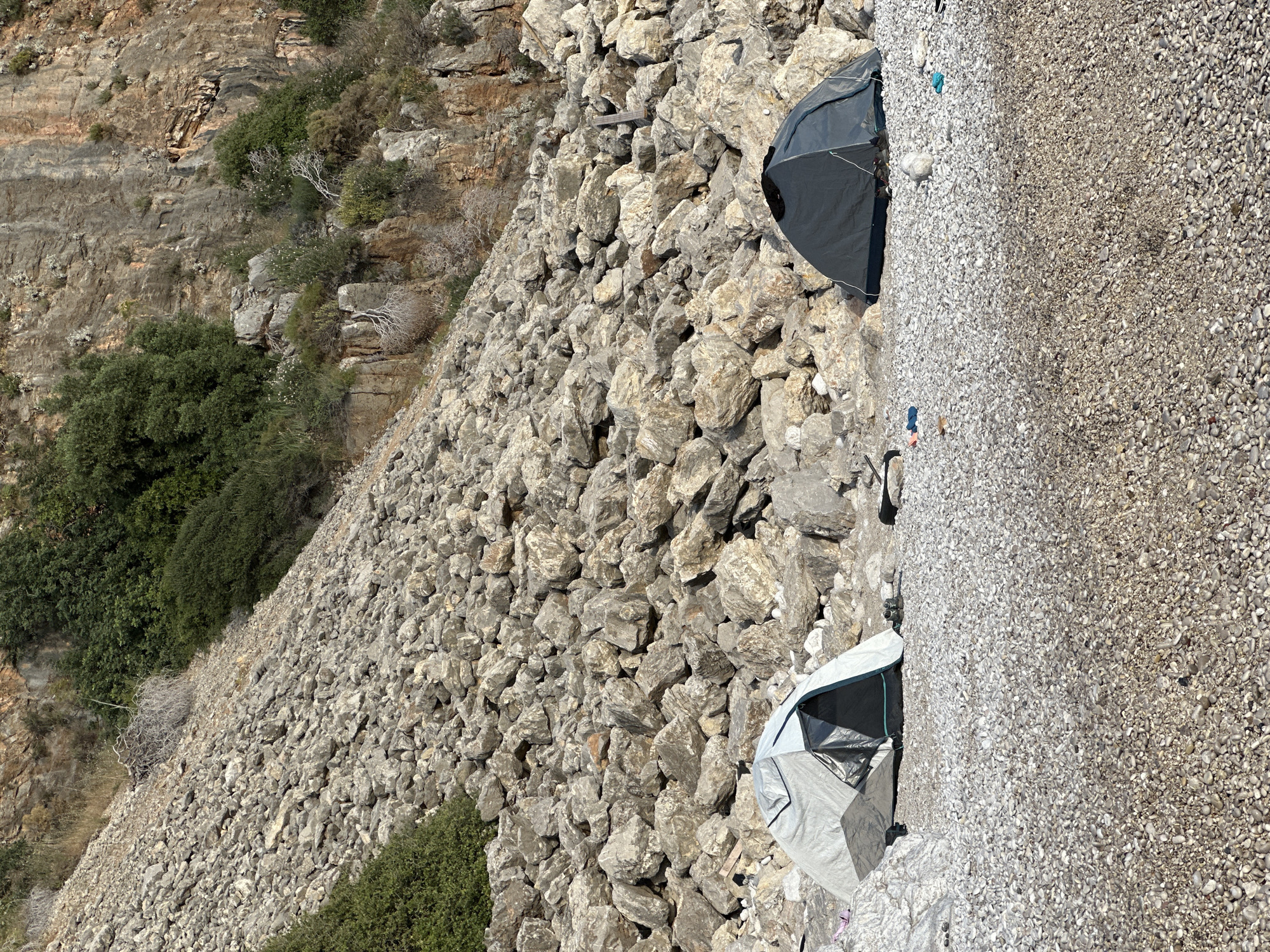 The image depicts a campsite located on a rocky beach in Fethiye, Turkey. Two tents are visible: a dark-colored one on the upper right and a light-colored one on the lower right. The area between the tents is covered with small, smooth stones. To the left of the tents, a steep slope composed of large rocks and boulders rises upwards, with sparse vegetation clinging to its side. In the background on the left is a weathered, rocky cliff face. No people are visible in the image. The scene appears to be a secluded camping spot in a rugged, coastal environment.