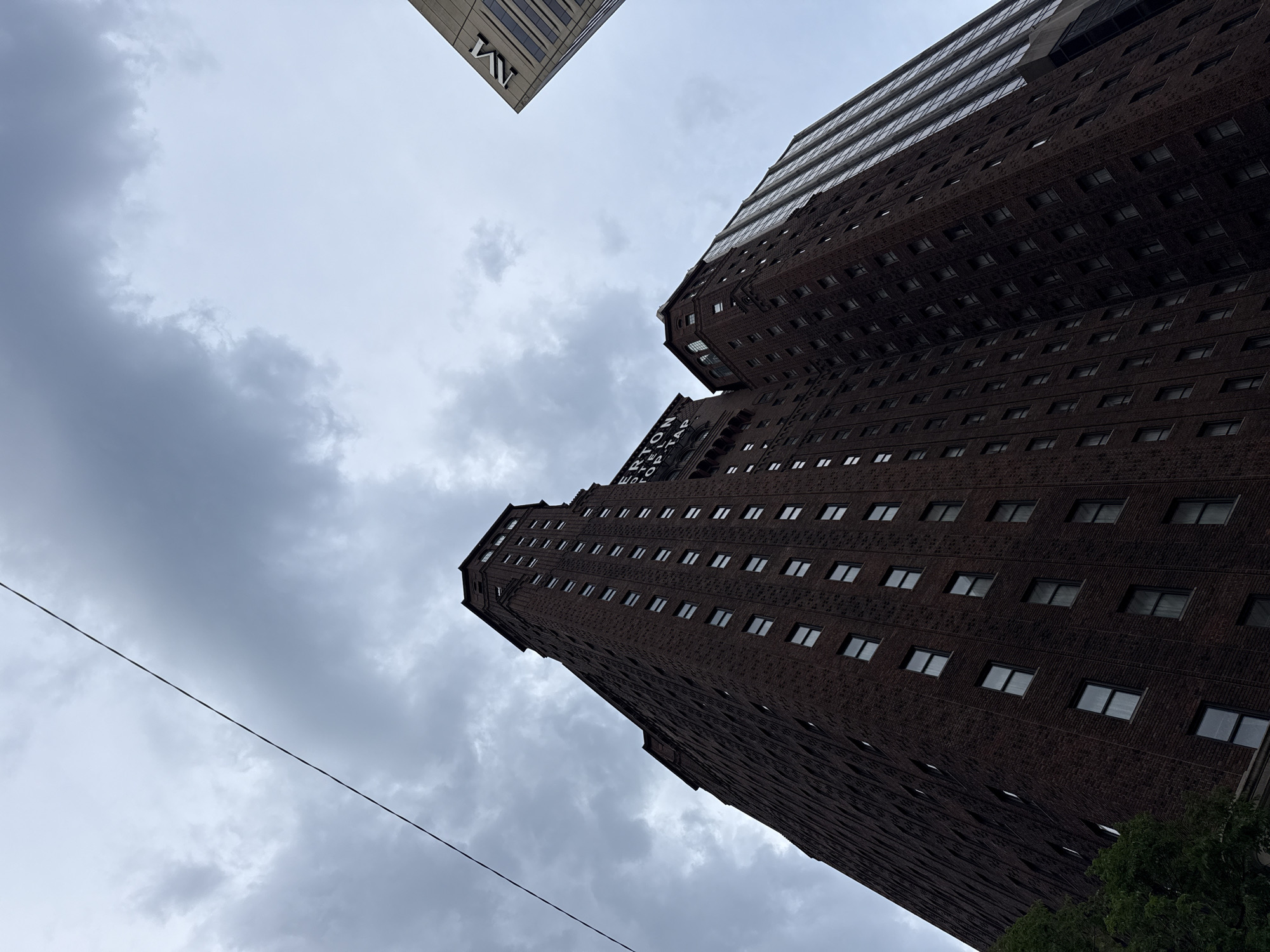 The image depicts a low-angle view of several tall buildings against a cloudy sky. The prominent structure on the left is a dark brick building with numerous windows, appearing to be a historic hotel. Text on the building is partially visible, reading "TERTON HOTEL" and "TOP TAP". To the right of this building, another tall structure with a glass facade is visible. In the upper right corner, a portion of a lighter-colored building with the "M" logo of a financial institution is seen. A thin dark line, possibly a wire or cable, bisects the upper left portion of the image. The scene suggests an urban environment, likely in Chicago, United States of America, given the architectural style and the presence of recognizable city elements. The lighting is diffused due to the overcast sky, creating a somewhat somber atmosphere. There are no people visible in the image.