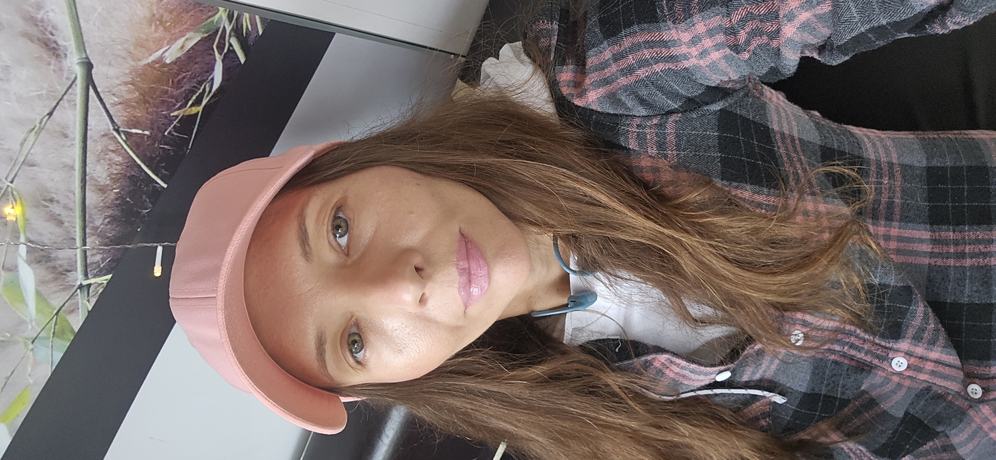 A medium shot shows a woman wearing a pink baseball cap, a white t-shirt, and a grey and pink plaid button-up shirt. She has long, brown hair, green eyes, and is wearing pink lip gloss. The woman is wearing a blue neckband around her neck. In the background, a framed picture or mural is visible, featuring bamboo stalks and what appears to be a panda. The lighting is bright, casting some shadows on the woman's face. The scene appears to be indoors. This image was taken in the city of Vitsebsk, Belarus.