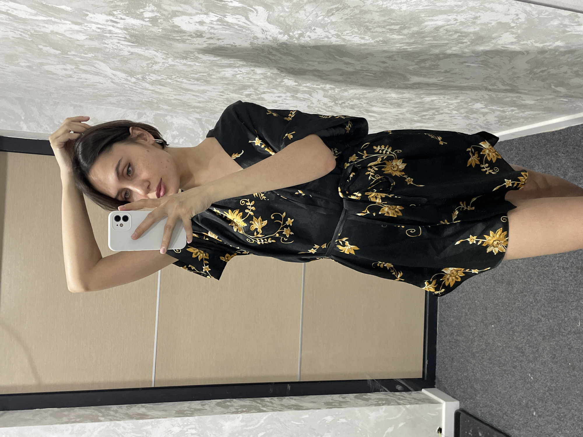 A young woman is taking a selfie in what appears to be an indoor setting. She is holding a white smartphone in her right hand, her left hand is touching her hair. She has short, dark hair and is wearing a black dress with yellow floral patterns.
The background includes a beige or light-colored wall, a white marble-like countertop, and a dark-colored frame or edge. The ceiling has a textured pattern resembling marble. The room appears to be a bathroom or dressing room. The photo was not taken in Izhevsk, Russia, there is no indication of the city in the photo.