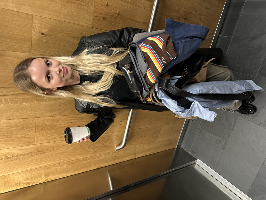 A blonde female, estimated to be in her 20s or 30s, stands centered in what appears to be an elevator or a confined room. She is dressed in a black leather jacket over a dark top and light-colored trousers, with black flat shoes visible. Her long blonde hair is parted in the middle. She holds a white disposable coffee cup, marked "Caramel," in her right hand. Her left arm supports a large bundle of clothing and accessories, including a brown and multi-colored striped Prada sweater, a light blue collared shirt, dark blue fabric, a grey garment, a dark leather belt, and black Prada shoes. She looks directly at the camera with a subtle smile. The immediate environment consists of light wood-paneled walls, a metallic handrail spanning the right background, and a reflective metallic panel on the left. The floor is composed of dark grey square tiles. The specific location within the city of London, United Kingdom, is not discernible from the visual information provided in the image.