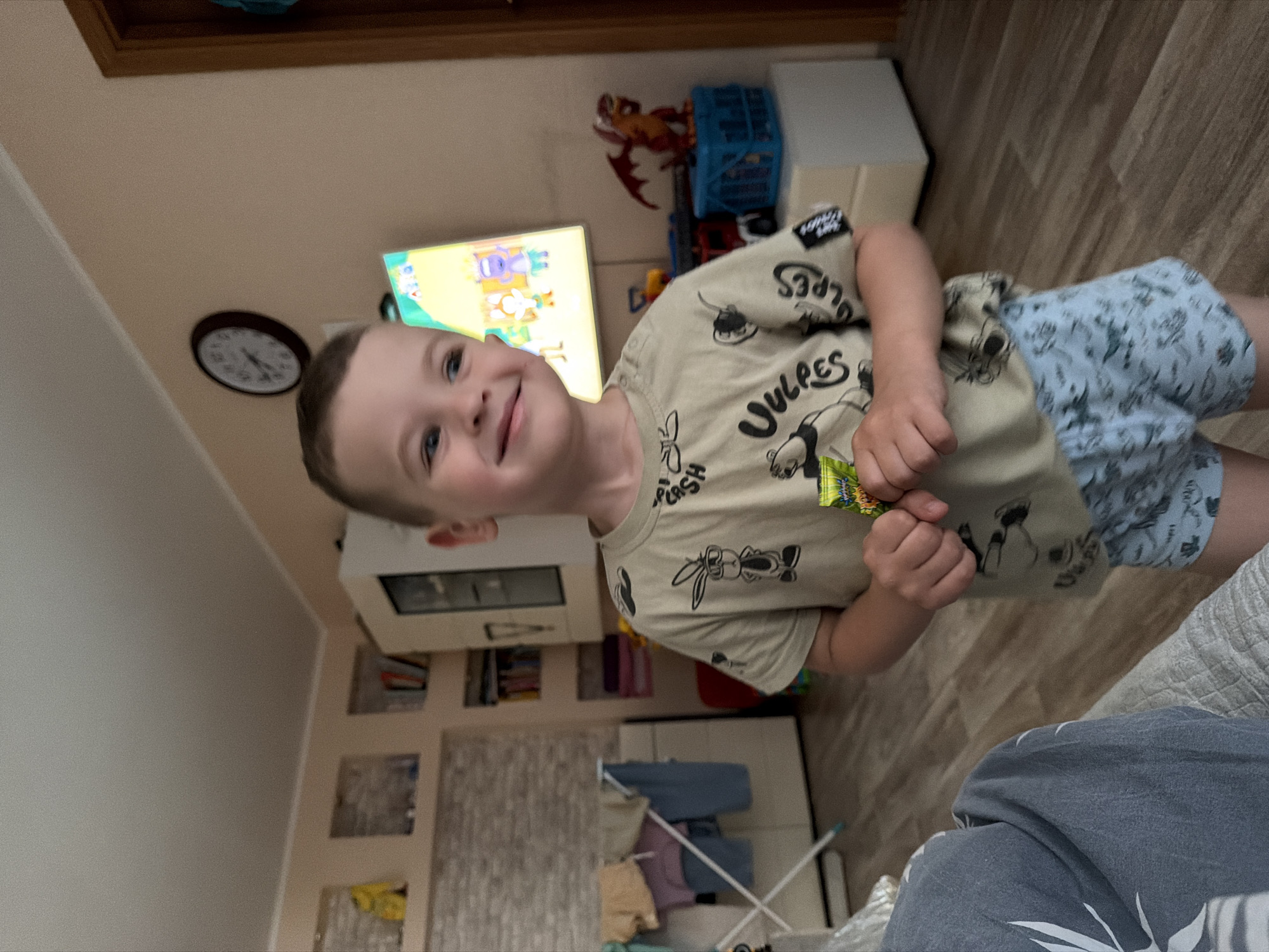 The image depicts a young boy standing in a room, holding a candy. The boy is wearing a tan t-shirt with cartoon characters and blue patterned shorts. In the background, a television displays a colorful cartoon, a clock hangs on the wall, and a small dragon figurine sits on a blue cage. Various furniture pieces, including shelves and a drying rack with clothes, are visible in the room. The overall setting appears to be a domestic interior. It is impossible to determine the specific location within the city of Khabarovsk, Russia, from the visual information provided.