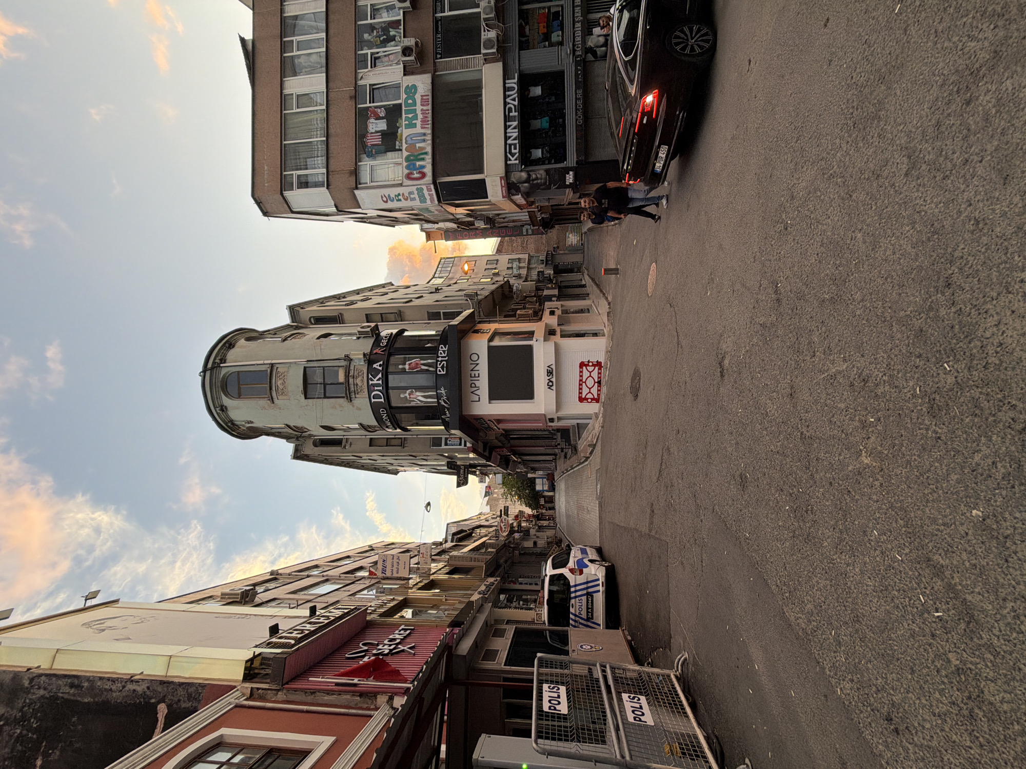 A street scene in Istanbul, Turkey, features a narrow street flanked by multi-story buildings on both sides. The sky above is partially cloudy with hues of orange and pink, indicating dusk. On the left, a white police vehicle with the word "POLIS" prominently displayed is parked behind a police barrier. The buildings on the left house various shops with signage including "JEREMY," "SECRET," and a red Turkish flag. The right side of the street showcases shops with signs such as "DIKA," "LAPİENO," "CEREN KIDS," and "KENN PAUL." Two men are walking on the right side of the street, approaching a dark-colored sedan parked further down the road. The street itself is paved and appears to be a pedestrianized area or a low-traffic street.