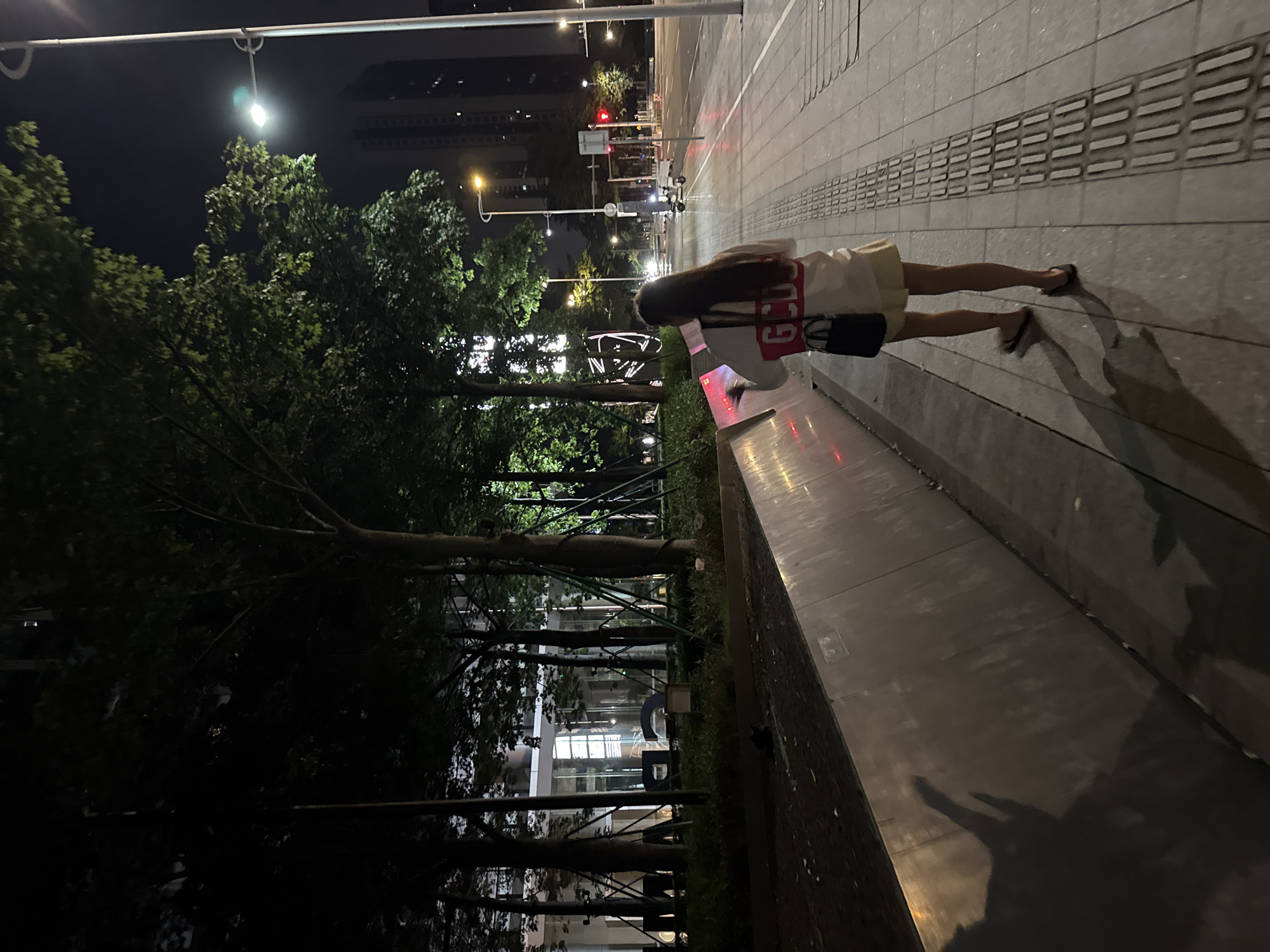 A medium shot captures a young woman walking at night on a well-lit sidewalk in the Haizhu District of China. She is facing away from the camera, her back to the viewer, and is interacting with a display panel. She wears a white t-shirt with red lettering that reads "GCD" across the back, a light yellow skirt, and flip-flops. A black handbag hangs from her right shoulder. Her shadow is cast prominently on a textured wall to her left. The environment includes trees with green leaves, a building in the background, streetlights, and a road with a vehicle visible in the distance. The sidewalk is paved with gray tiles and features a tactile paving strip. The scene suggests an urban setting at night.