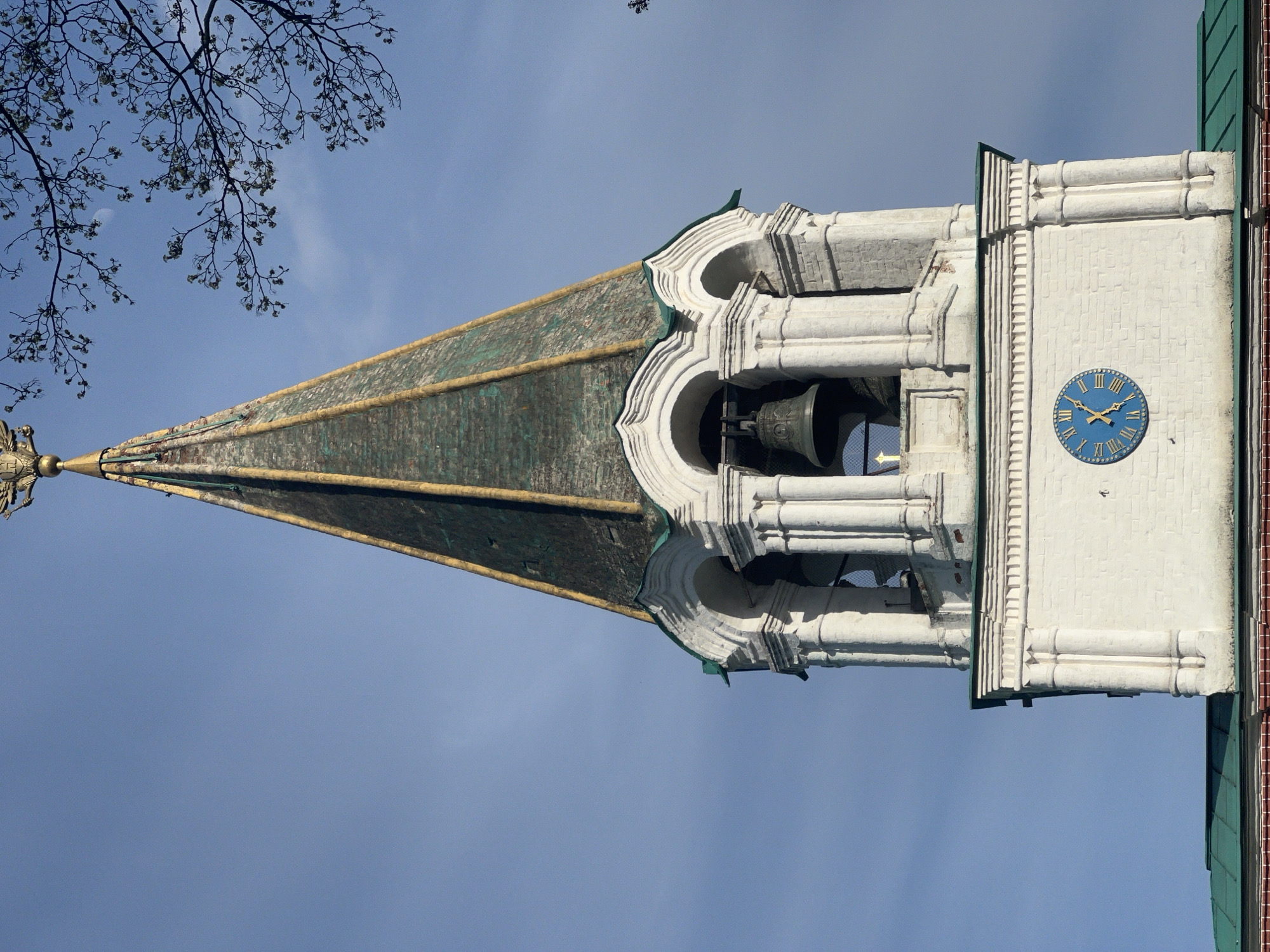 The image shows the Ivan the Great Bell Tower, located in the Moscow Kremlin, Moscow, Russia. It features a tall, green-patina roof structure with golden trim rising to a point and topped with a cross. The tower below the roof is white with arched openings containing bells, and a clock face with blue background and Roman numerals is visible on the side. Some bare tree branches are in the upper left corner of the frame, and the sky is a clear, pale blue.