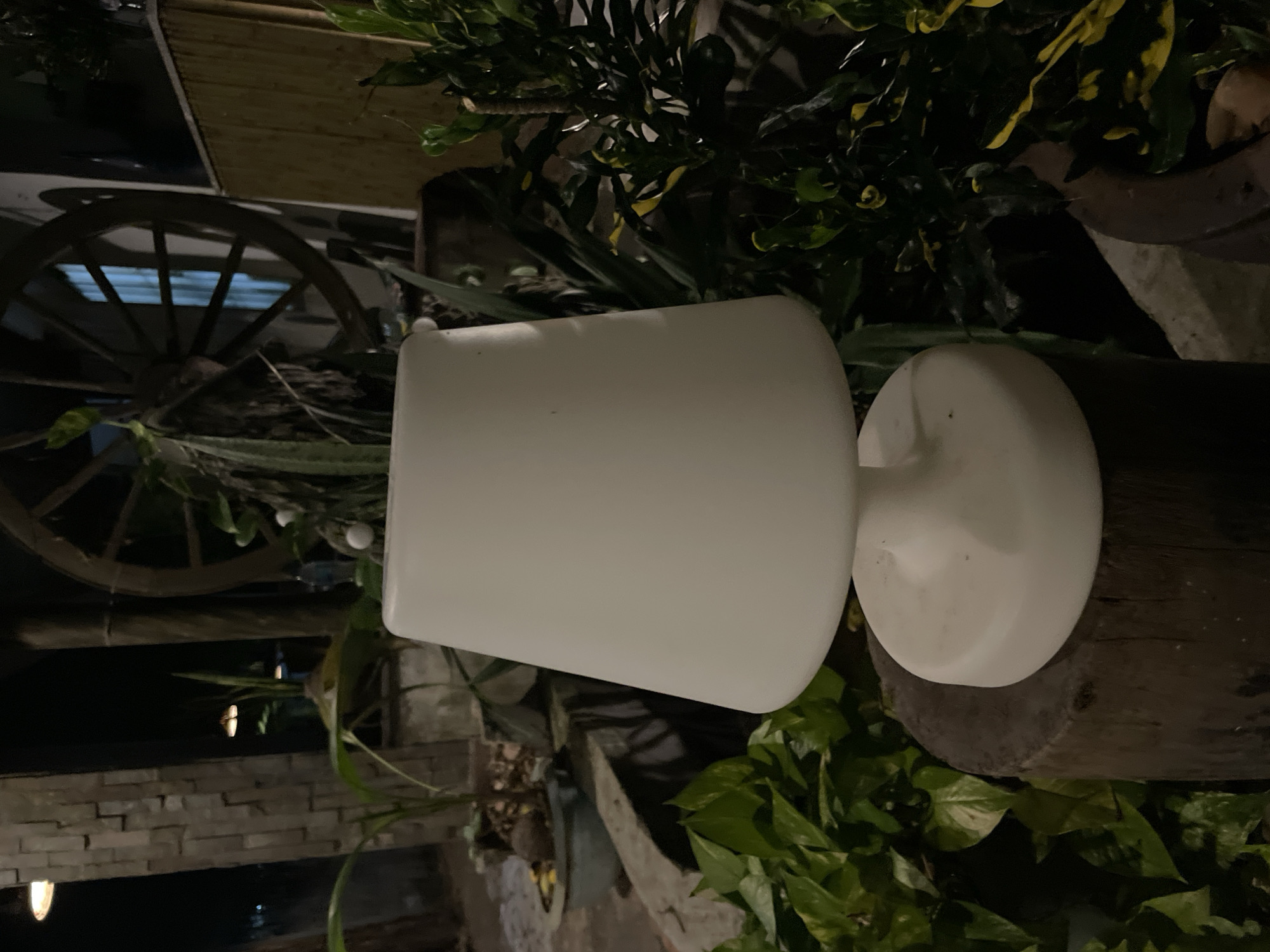 A white lamp sits on a wooden stump in an outdoor area at night. Behind the lamp, there are plants, a stone wall, and a large wooden wheel. The area appears to be a courtyard or garden within a property in Phu Thuy Ward, Vietnam.