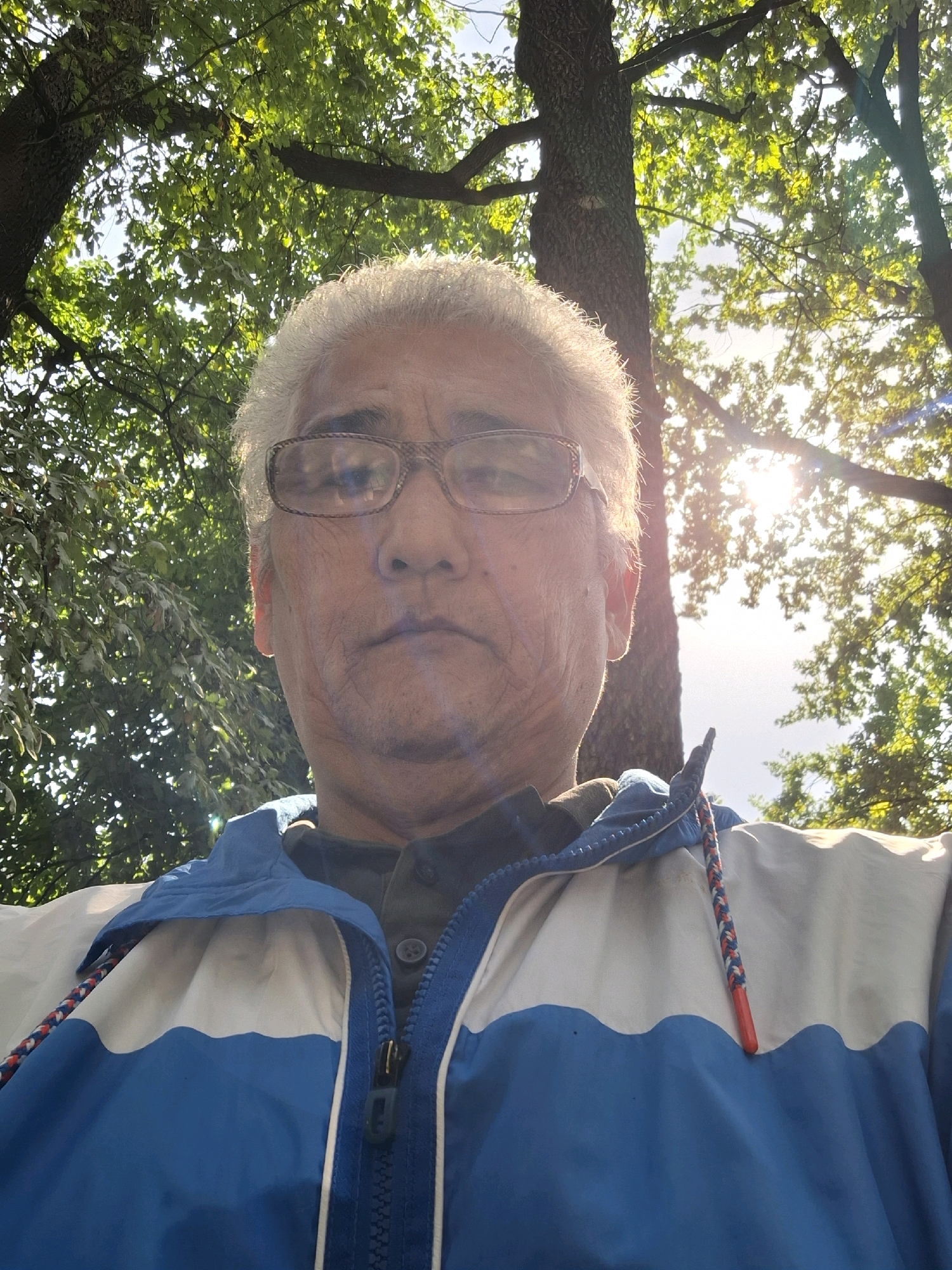 The image is a selfie of an elderly Asian man with white hair, wearing glasses, and a blue and white jacket. He is outdoors, with trees and sunlight in the background. The location is not specified in the image.