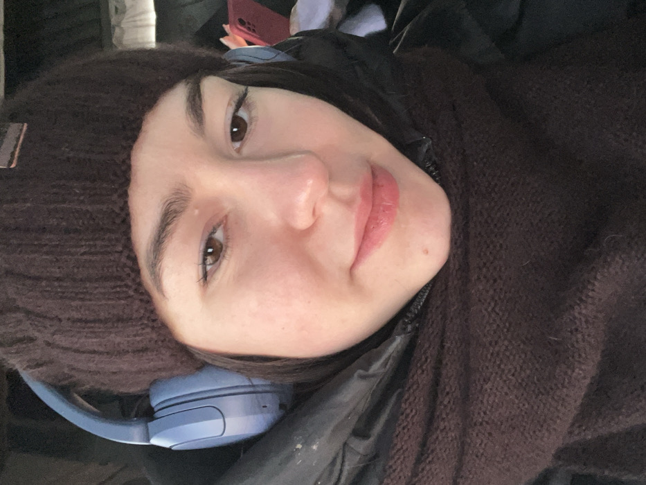 A young adult female subject is depicted in a close-up, selfie-style photograph taken in the city of Pyriatyn, Ukraine. She is wearing a dark brown knit beanie and light blue over-ear headphones. Her attire also includes a dark-colored puffer jacket and a dark brown knit scarf wrapped around her neck, covering the lower portion of her face. She has brown eyes, a slight smile, and is wearing light pink lipstick. The subject is looking directly towards the camera. The background is blurred and indistinct, suggesting an indoor or confined environment.